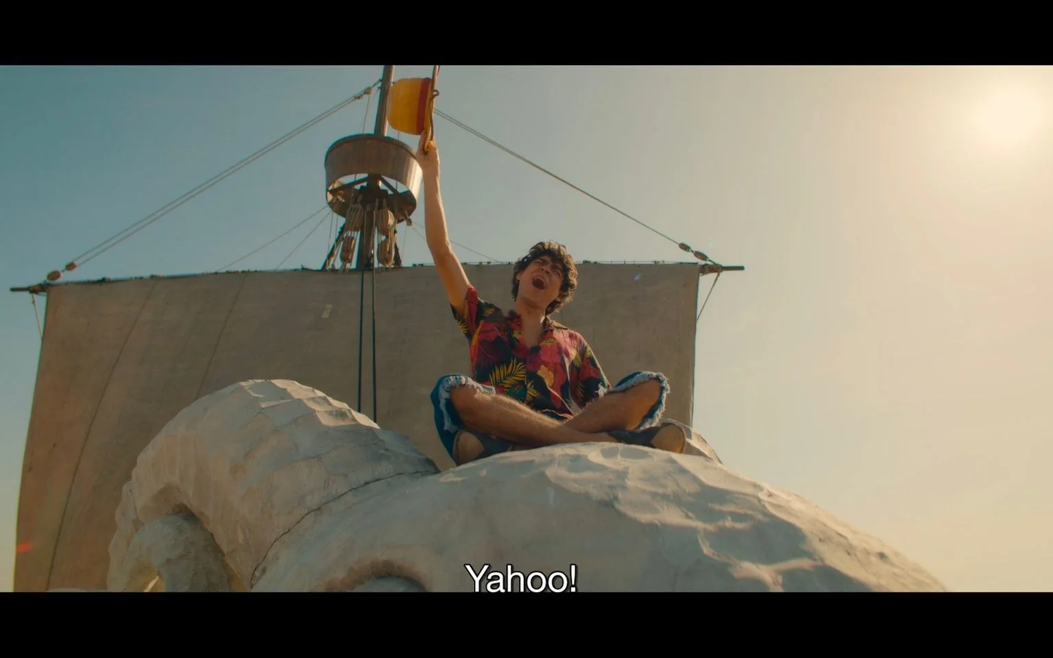 The image shows a character named Luffy from "One Piece" sitting atop a large, stone-like structure that resembles a sheep's head on a ship. Luffy is wearing a vibrant, multicolored, tropical shirt and ripped denim shorts, exuding a carefree and adventurous spirit. He is energetically raising a straw hat in the air, expressing joy and excitement, as indicated by the subtitle "Yahoo!" The ship's large sail is visible behind him, casting a soft shadow and contributing to the nautical theme. The sun shines brightly in the clear sky, highlighting the cheerful and exuberant mood of the scene. The composition captures a sense of freedom and adventure, with Luffy's pose suggesting a triumphant and carefree demeanor.