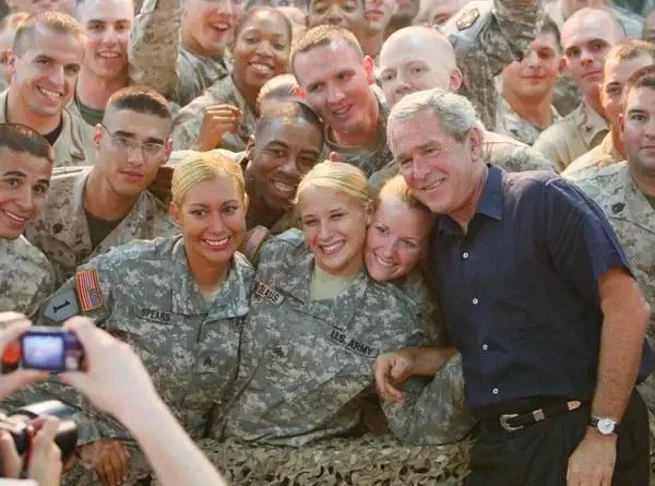 President George W. Bush is pictured visiting troops in Iraq, surrounded by a group of smiling soldiers. The soldiers are in military uniforms, with visible name tags and insignias on their chests and sleeves. Bush is positioned on the right side of the image, wearing a dark short-sleeved shirt and smiling as he poses with the soldiers. The atmosphere is lively and positive, with the soldiers appearing enthusiastic and engaged. The setting appears to be indoors, possibly in a tent or military facility, suggested by the camouflage netting in the foreground. The lighting is warm, illuminating the faces of the group. The soldiers have different expressions ranging from cheerful to proudly serious, displaying camaraderie and appreciation for the visit. A camera is visible in the foreground, capturing the moment as others gather closely for the photo opportunity.