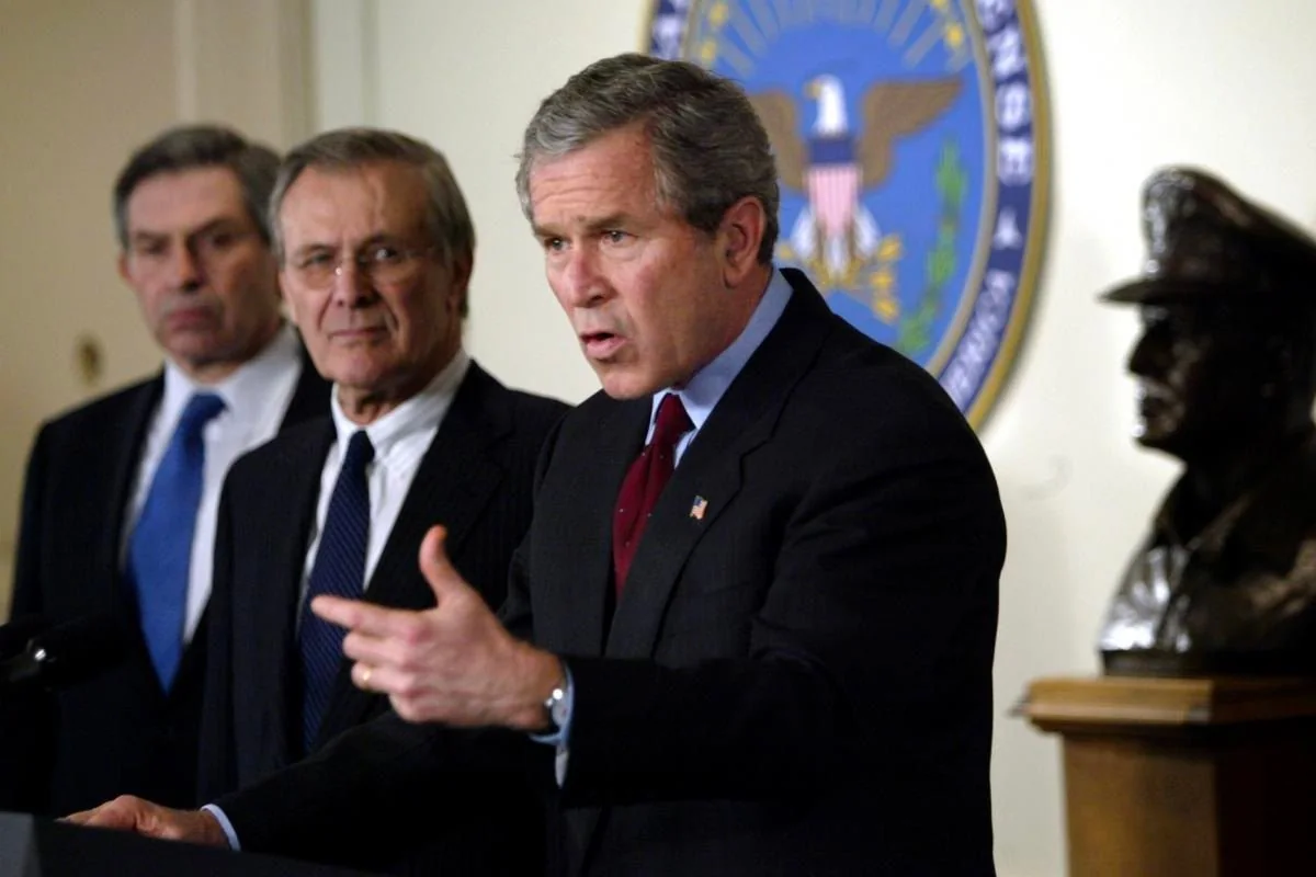 In this indoor scene, President George W. Bush is standing at a podium, delivering a speech or making an important announcement. He is wearing a dark suit with a red tie and a white shirt, exuding a formal and authoritative presence. His expression is serious and focused, emphasizing the gravity of the occasion. In the background, two men are visible, each dressed in dark suits and ties, listening attentively. The backdrop features a large emblem with an eagle, likely representing a government or military department, adding to the official atmosphere. A bust of a military figure is also visible on a wooden stand to the right, further reinforcing the governmental setting. The lighting is bright and evenly distributed, highlighting the individuals and the emblem behind them, creating a clear and professional image suitable for a press conference or official statement.