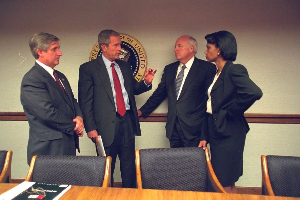 The image depicts a formal meeting setting with four individuals engaged in discussion. In the center, President George W. Bush is gesturing with his hand, appearing to explain or make a point. To his right stands Condi, likely Condoleezza Rice, listening intently with her hands on her hips, dressed in a dark suit with a white blouse. To the left of President Bush, another man is dressed in a suit with a maroon tie, his hands resting in front of him, appearing attentive. Further to the right, another man in a suit and polka dot tie listens with a serious expression.
The background features the seal of the President of the United States, adding a formal and official tone to the setting. The room is equipped with a wooden table and chairs, suggesting a meeting or conference room atmosphere. The lighting is bright and even, typical of an indoor office environment. The overall mood appears professional and focused, indicative of a serious discussion or briefing.