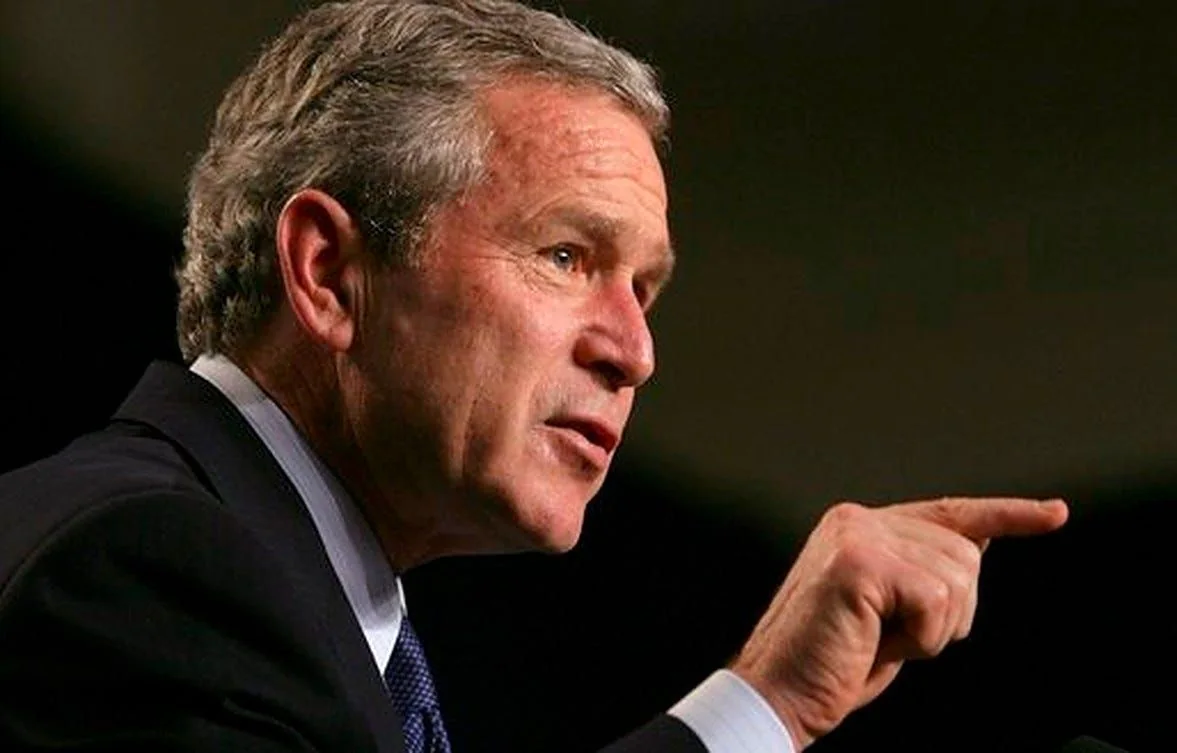 President George W. Bush is depicted in a dynamic profile view, emphasizing a moment of animated speech or discussion. The lighting is focused, highlighting his facial features and enhancing the serious tone of the scene. He is wearing a dark suit with a white shirt and a subtly patterned tie, projecting a formal and authoritative presence. His expression is intense and focused, with his mouth slightly open as if mid-sentence, and his eyes are directed towards something or someone out of the frame, suggesting engagement with an audience or interlocutor. The background is dark and unobtrusive, ensuring full attention remains on President Bush. The image captures a sense of immediacy and importance, likely taken during a public address or press conference. The positioning of his hand, with a pointed finger, adds to the impression of emphasis or persuasion in his speech. The overall color palette is subdued, reflecting the seriousness of the occasion.
