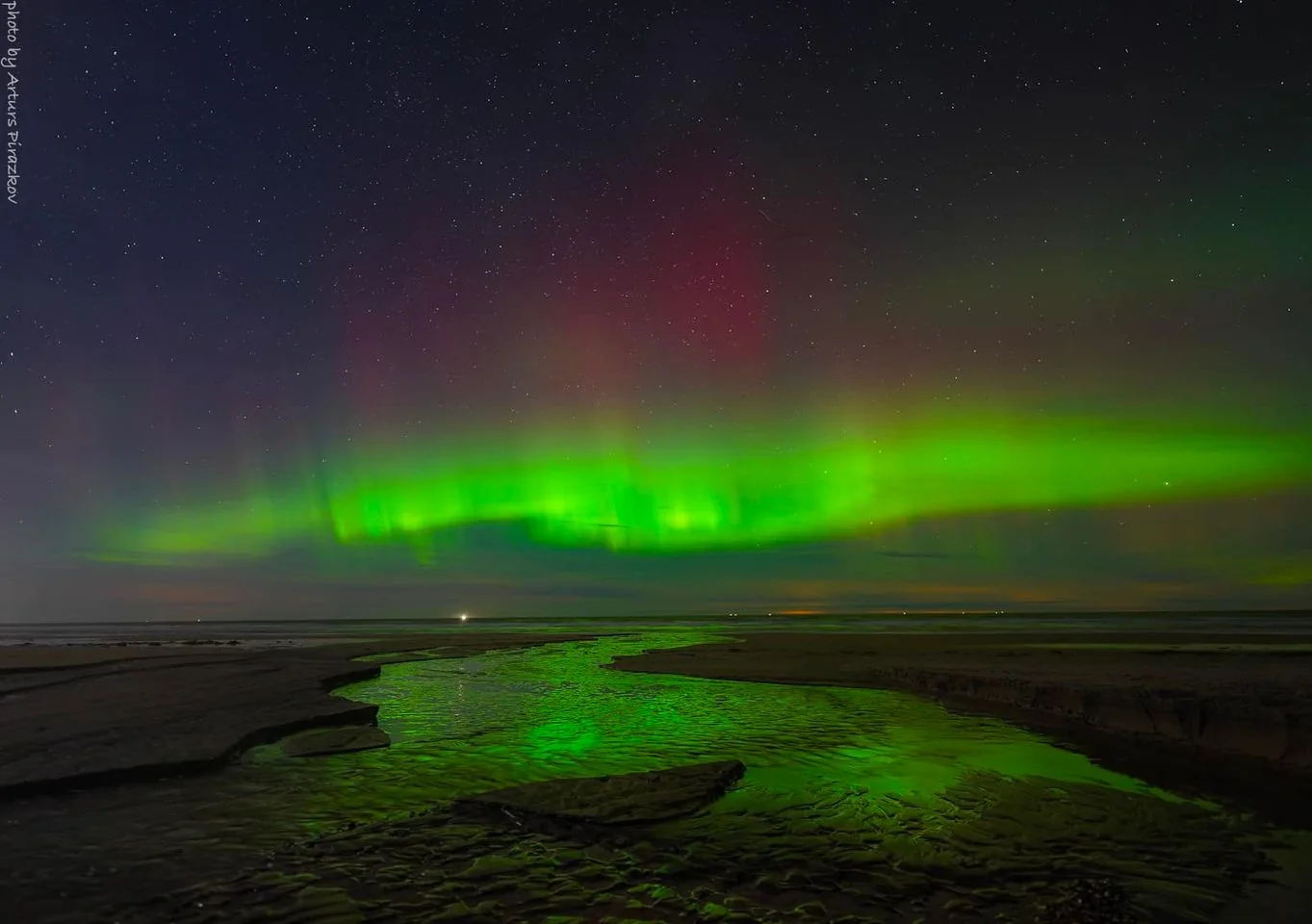 The image captures a stunning view of the northern lights, or aurora borealis, stretching across the night sky. Vibrant green and subtle red hues curve and dance across the horizon, creating a mesmerizing display of natural beauty. The night sky is dotted with countless stars, adding to the ethereal atmosphere of the scene. In the foreground, a winding stream reflects the vibrant colors of the aurora, enhancing the visual impact of the scene. The landscape is serene and expansive, with the land gently sloping toward the horizon. The lighting is soft and ambient, with the natural glow of the aurora providing the primary illumination. The overall mood is tranquil and awe-inspiring, capturing a moment of serene beauty in nature. The photograph is credited to Artem Panteleev, as indicated by the on-screen text.