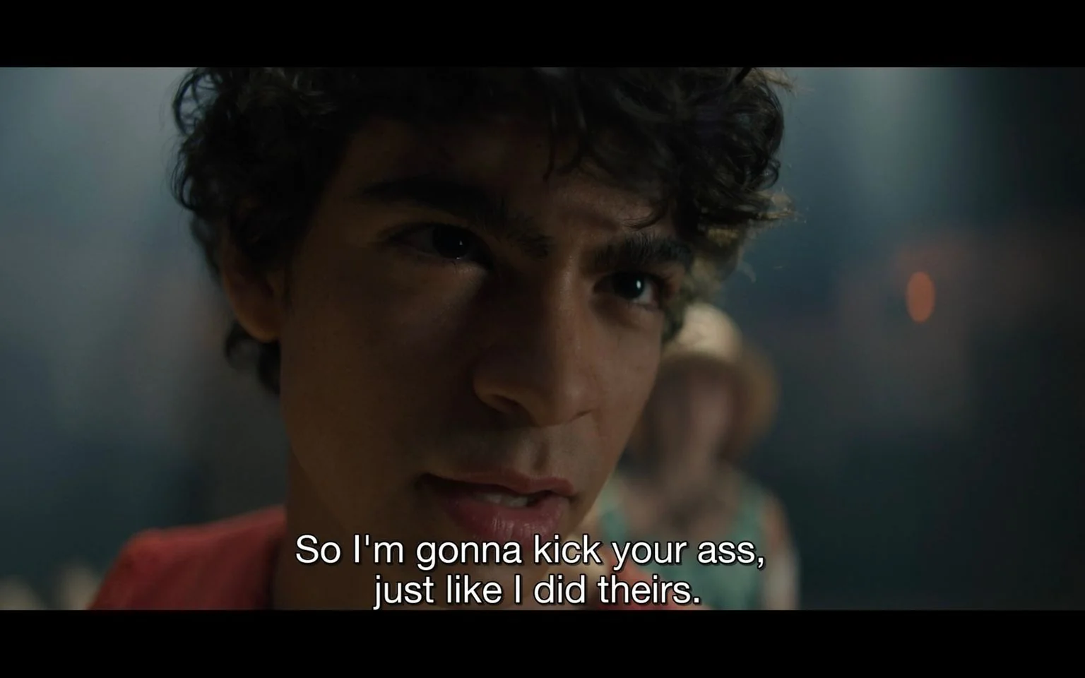 The image is a close-up of a young man with curly hair, delivering a confident and intense expression. The lighting is dim, highlighting the serious mood of the scene. The background is blurred, suggesting a focus on the character's facial expression and words rather than the setting. He is speaking with determination, indicated by the subtitle: "So I'm gonna kick your ass, just like I did theirs." This suggests a confrontational or competitive context. The person is wearing a red shirt, adding to the bold and assertive tone. The atmosphere is tense, with a spotlight effect that emphasizes the character's seriousness and focus.