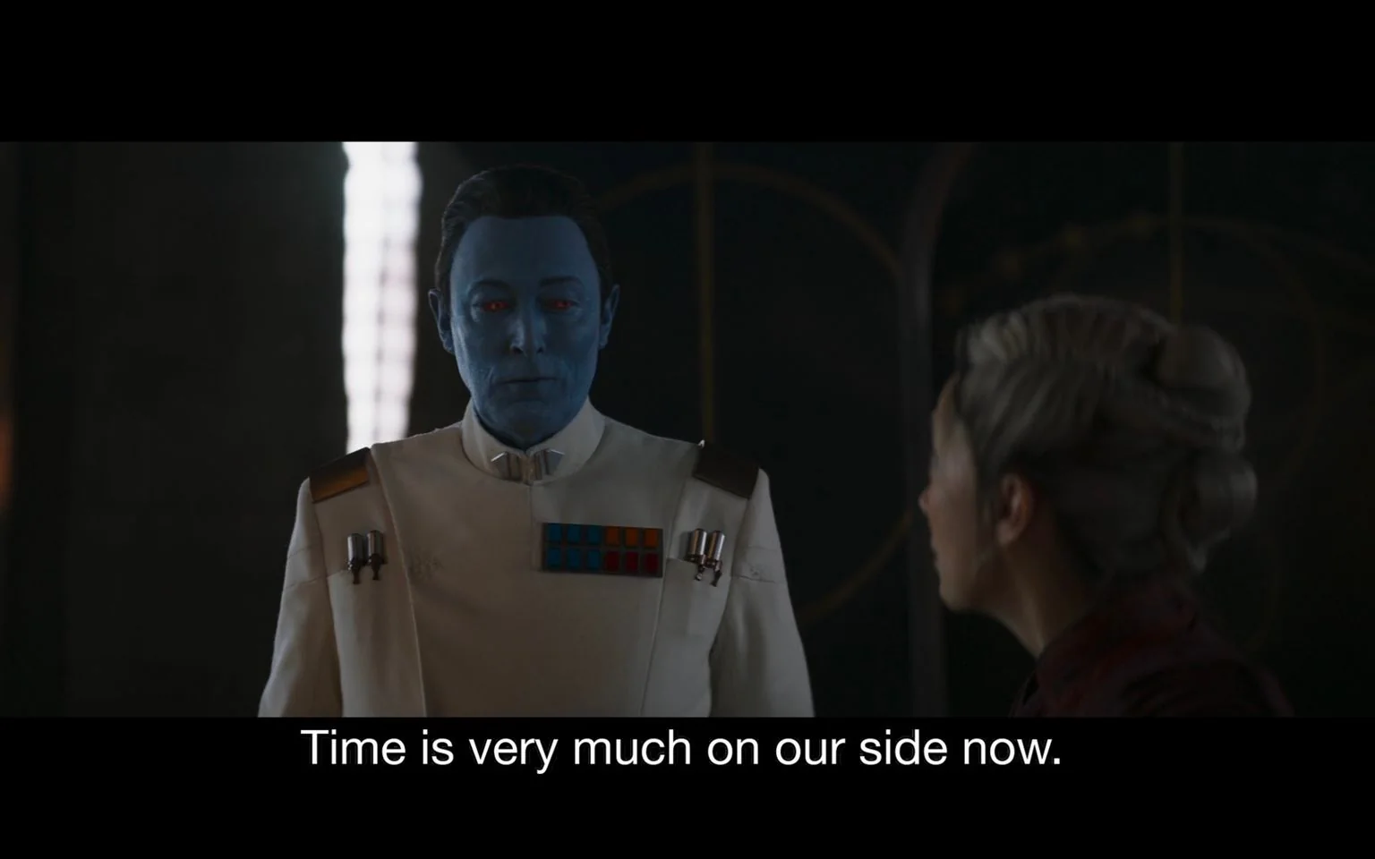 The image is a scene from a "Star Wars" production. It features a blue-skinned character wearing a white military uniform adorned with colorful insignia and epaulets, indicating a high rank. His expression is calm and composed as he engages in a conversation. He has red eyes and is standing in a dimly lit room, with another person visible from behind in the foreground. The other person is facing him and has intricately styled hair. The setting appears to be an interior space with a dark, possibly metallic or stone background, adding to the serious and strategic atmosphere of the scene. The lighting highlights the characters, creating a stark contrast with the darker background. The caption at the bottom states, "Time is very much on our side now," suggesting a strategic or tactical discussion.