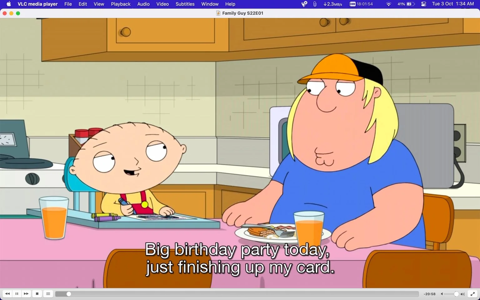 ```
The image is a screenshot from an episode of the animated television show "Family Guy" (Season 22, Episode 1), viewed on VLC media player. In the scene, two characters, Stewie and Chris, are sitting at a kitchen table. Stewie, a baby with a distinctive football-shaped head, is on the left, wearing his usual yellow and red outfit. He appears to be drawing or writing on a card, with a glass of orange juice and crayons visible in front of him. Chris, a teenager with long blond hair and wearing a blue shirt and an orange cap, is on the right, eating a meal with a glass of juice. The kitchen setting is warm and homey, with wooden cabinets and a backsplash visible in the background. The subtitle reads, "Big birthday party today, just finishing up my card," indicating a casual conversation between the two. The animation style is typical of the show, with bold outlines and bright colors.