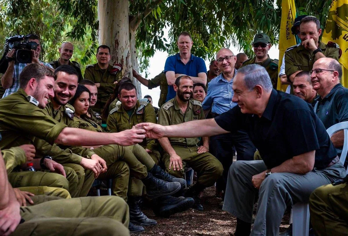 In this outdoor scene, Benyamin Netanyahu is interacting with a group of people in military uniforms. The setting appears casual, with everyone seated under trees, creating a relaxed atmosphere. Netanyahu, wearing a black shirt and light-colored pants, is leaning forward from his chair, reaching out to shake hands with a person in the group, suggesting a gesture of camaraderie or thanks.
Around Netanyahu, several individuals in military attire are seated in a semicircle, some smiling, indicating a positive mood. The group includes both men and women, and their uniforms with various insignias suggest they are part of a military organization. In the background, a few more individuals can be seen standing, observing the interaction, and a photographer captures the moment, adding to the sense of a significant visit.
The colors are natural, with the green of the uniforms blending with the foliage around. The lighting suggests a daytime setting, enhancing the overall sense of approachability and engagement. The composition focuses on the interaction between Netanyahu and the group, highlighting the connection and rapport being shared.