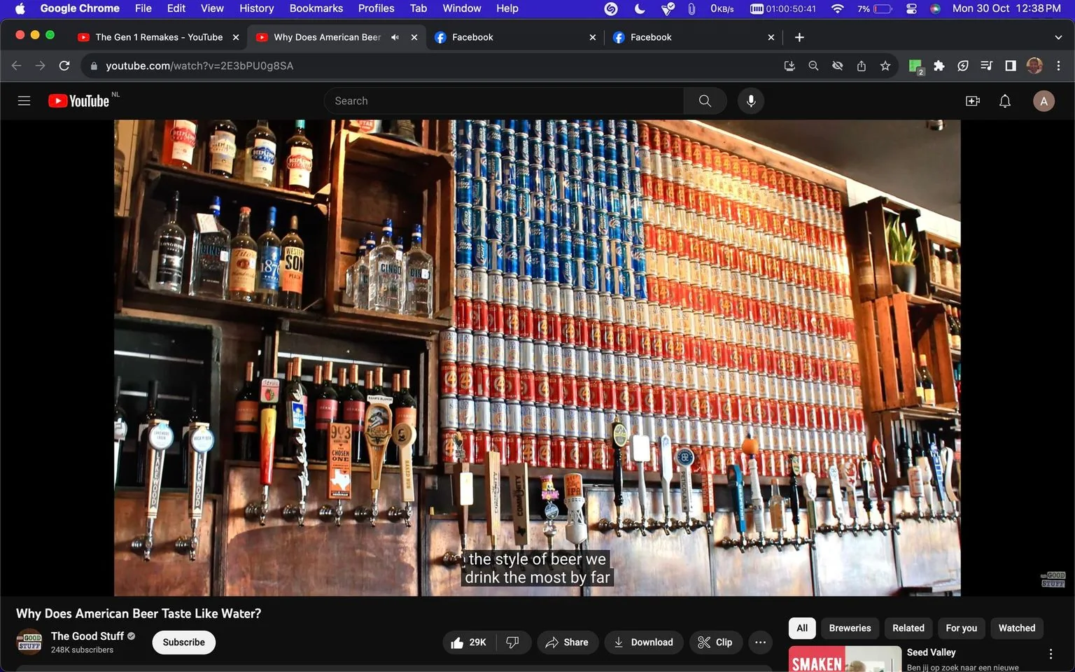 The image is a screenshot from a YouTube video titled "Why Does American Beer Taste Like Water?" on the channel "The Good Stuff." The scene captures the interior of a bar, featuring a prominent display of beer taps and shelves stocked with various liquors. A striking visual element is the American flag made from stacked beer cans in red, white, and blue, occupying a large portion of the wall behind the taps. Several liquor bottles and wine bottles are visible on wooden shelves, adding to the bar ambiance. The lighting is warm, highlighting the rustic and inviting atmosphere. The subtitles at the bottom read, "the style of beer we drink the most by far," suggesting a discussion about popular beer styles. The overall setting conveys a traditional and patriotic American bar environment, emphasizing the theme of American beer.