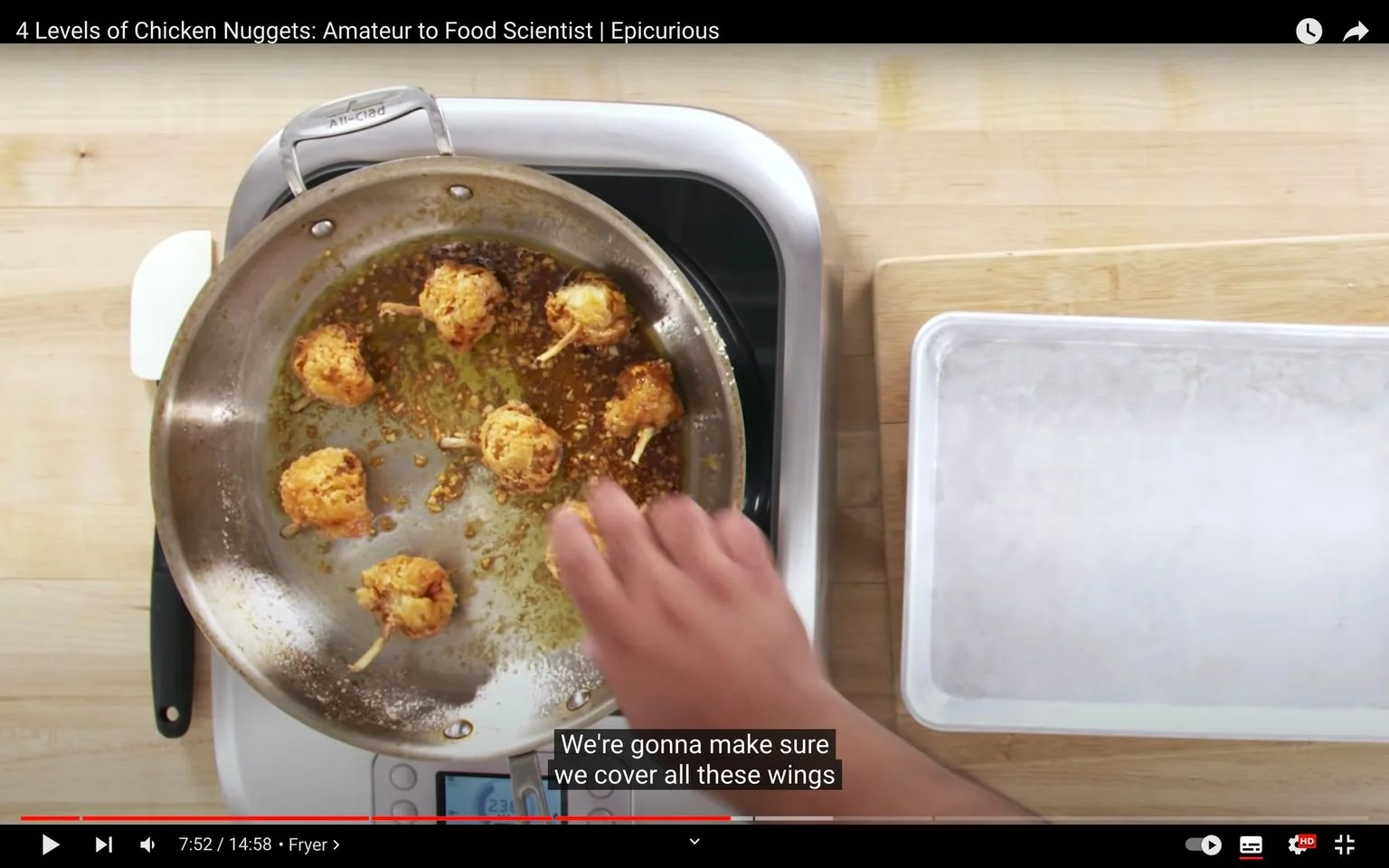 1) Rich description: The video frame is from a YouTube video titled "4 Levels of Chicken Nuggets: Amateur to Food Scientist | Epicurious," published by Epicurious. It captures a cooking demonstration in a kitchen setting. A stainless steel pan is on a stovetop, containing several golden-brown, fried chicken pieces, each with a skewer. The pan appears to have remnants of fried bits and oil, indicating the cooking process is well underway. A person’s hand, partially visible, is interacting with the food, likely turning or coating the chicken pieces. The background includes a wooden countertop with a cutting board and an empty metal tray on the side, suggesting preparation and organization for the cooking process. The lighting is bright, focusing on the food and hands-on action in the pan. The scene conveys a practical and instructional atmosphere, typical of cooking tutorial videos.
2) On-screen text block:
- Title: "4 Levels of Chicken Nuggets: Amateur to Food Scientist | Epicurious"
- Site / app: "YouTube"
- Captions / subtitles: "We're gonna make sure we cover all these wings"