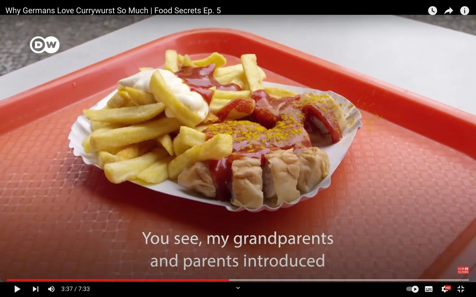 This video frame is from the YouTube video titled "Why Germans Love Currywurst So Much | Food Secrets Ep. 5," published by DW. The scene features a classic German dish, currywurst, served on a red tray. The currywurst is neatly arranged on a paper plate and topped with ketchup and curry powder, giving it a vibrant red and yellow appearance. Next to the sausage, a generous portion of fries is visible, some of which are topped with mayonnaise. The composition is simple yet colorful, emphasizing the appeal of the meal. The background appears to be an outdoor setting, typical for enjoying street food. The subtitle on the screen reads, "You see, my grandparents and parents introduced," suggesting a narrative about family and tradition connected to the dish. The overall atmosphere conveys a sense of cultural exploration, highlighting the significance of currywurst in German food culture.
- Title: "Why Germans Love Currywurst So Much | Food Secrets Ep. 5"
- Channel / profile: "DW"
- Site / app: "YouTube"
- Captions / subtitles: "You see, my grandparents and parents introduced"