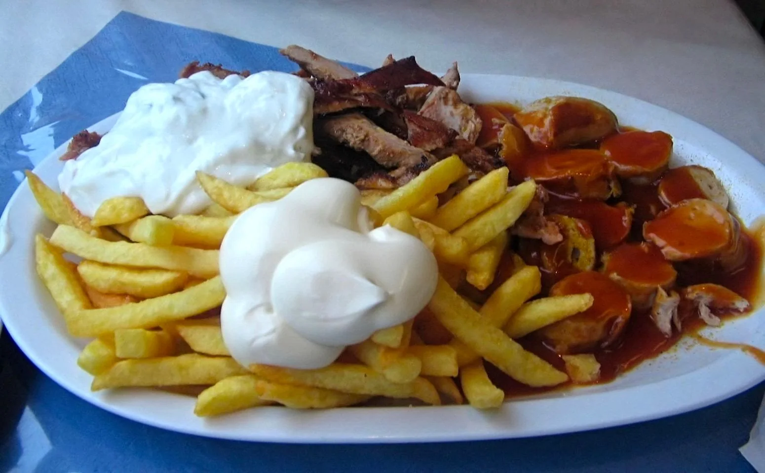 This image features a plate of food with a variety of items. On the plate, there are crispy-looking French fries topped with a generous dollop of white sauce, likely sour cream or mayonnaise. To the side, slices of grilled meat are visible, possibly gyro or doner kebab, accompanied by another portion of fries. Additionally, there is a serving of sliced sausages covered in a rich, reddish-brown sauce, possibly currywurst. The meal is presented on a white oval plate placed on a blue surface, which adds a cool contrast to the warm tones of the food. The overall presentation suggests a hearty, indulgent meal, typical of street food or casual dining. The lighting is natural, highlighting the textures and colors of the dish.