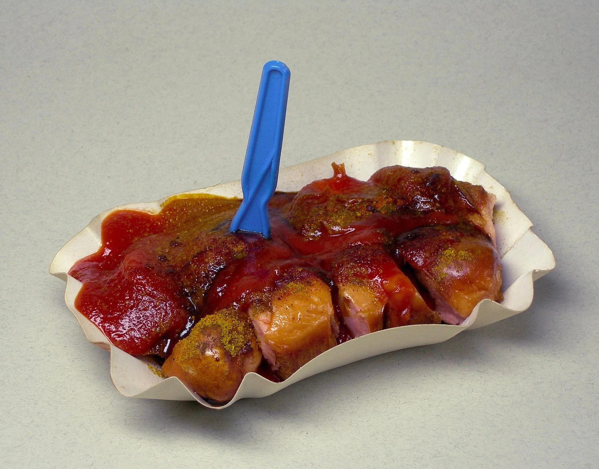 This image features a serving of currywurst, a popular German fast food dish. The sliced sausage is generously topped with curry ketchup and sprinkled with curry powder. It's served in a disposable paper tray, typical for street food or quick takeaway meals. A blue plastic fork is stuck into the sausage, ready for eating. The colors are vibrant, with the deep red of the sauce contrasting with the lighter shades of the sausage and the soft yellow of the curry powder. The lighting is bright and even, highlighting the glossy texture of the sauce. The plain background keeps the focus on the dish itself, emphasizing its simplicity and appeal as a savory snack.