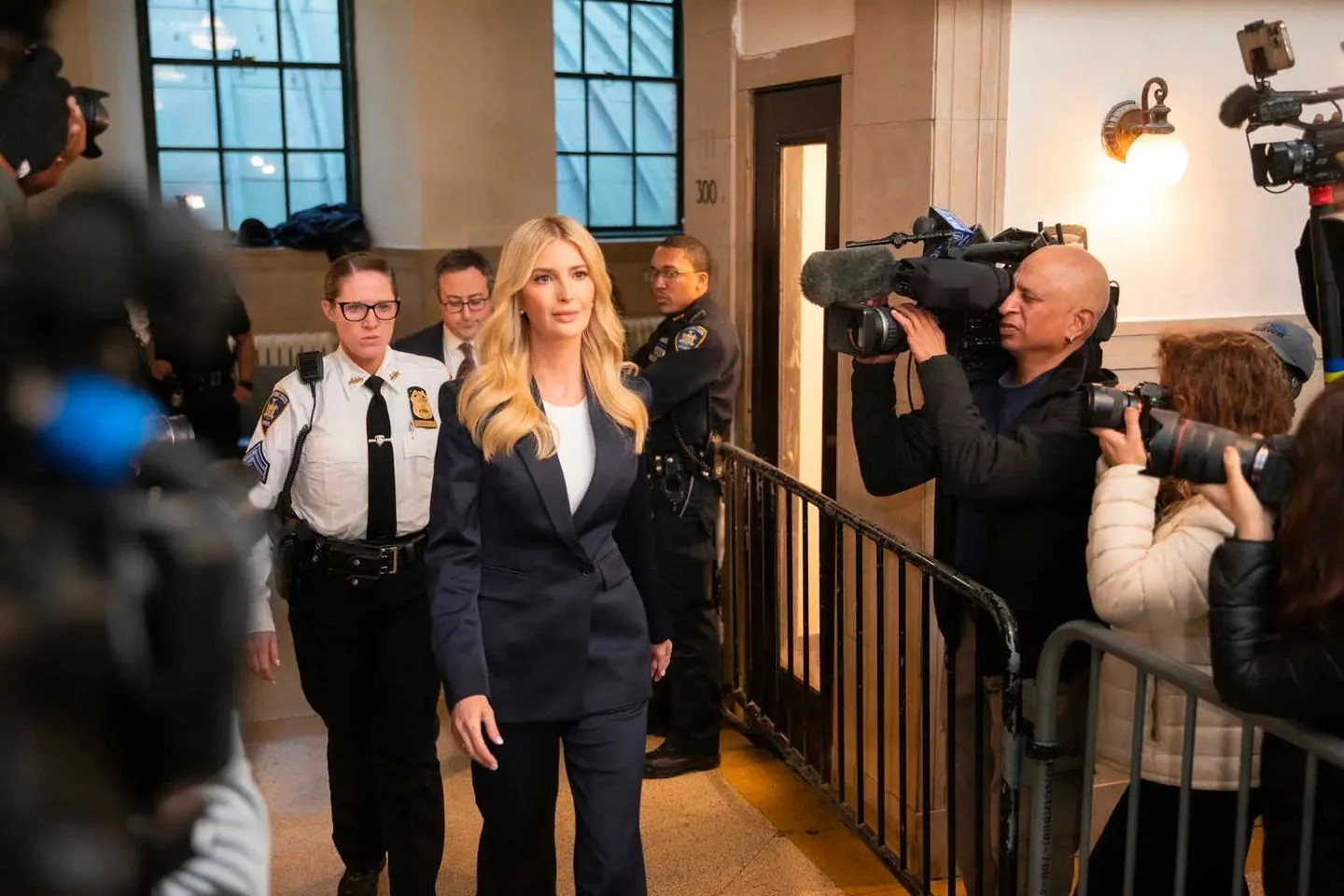 ```
Miss Ivanka Trump is seen walking in a formal setting, accompanied by security personnel. She is dressed in a tailored navy blue suit with a white top underneath. Her blonde hair is styled in loose waves. The atmosphere is busy, with multiple cameras and photographers capturing the moment. A police officer and several other security staff are visible, ensuring her safety. The setting appears to be indoors, possibly at a courthouse or similar official venue, indicated by the presence of security and the number "300" visible on a door behind her. The lighting is bright, emphasizing the professional and serious tone of the event. Photographers are actively documenting the scene, with some holding large cameras equipped with microphones and long lenses.
- Title: "..."
- Channel / profile: "..."
- Site / app: "..."
