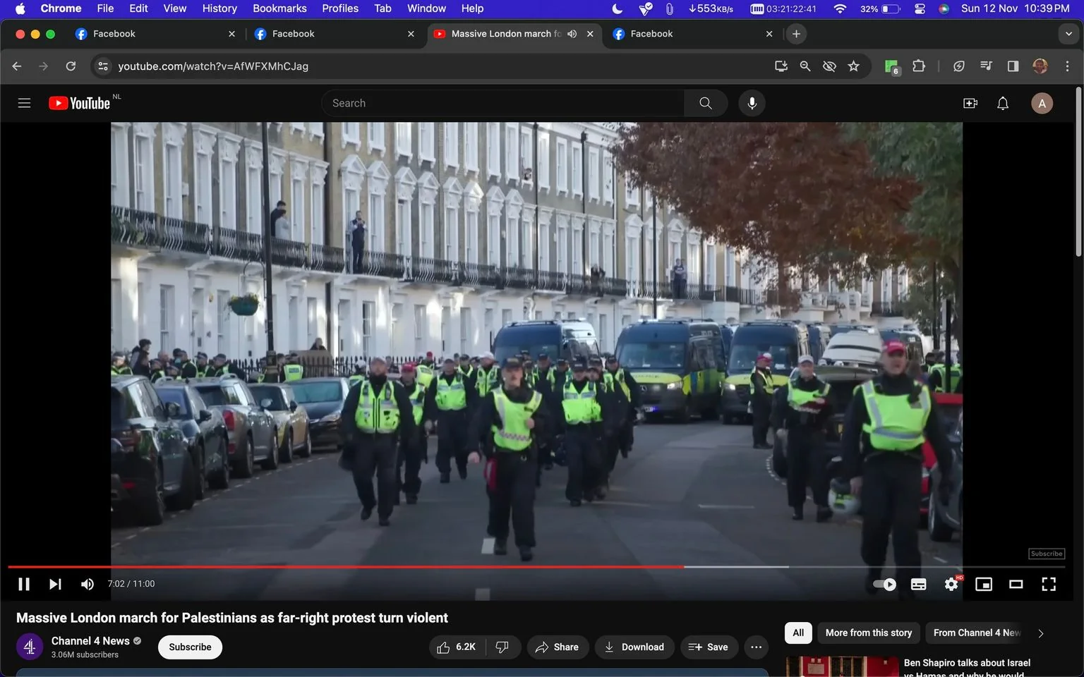 A screenshot from a YouTube video titled "Massive London march for Palestinians as far-right protest turn violent" is shown. This video is from the Channel 4 News channel. The scene captured is outdoors on a city street, likely in London, given the context. Rows of uniformed police officers, wearing black and neon yellow jackets, are walking down the street. The officers are in a coordinated formation, suggesting they are managing or responding to a public event, possibly a protest.
In the background, there are several police vans parked along the street, lined with typical London terraced houses. People can be seen observing from the balconies, indicating an event of significant public interest. The lighting suggests it is daytime, and the autumn foliage hints at a time around November. The visual composition emphasizes the large police presence, suggesting a significant or potentially volatile situation. The screenshot also shows the YouTube interface, with options like play, save, and download visible on the screen.