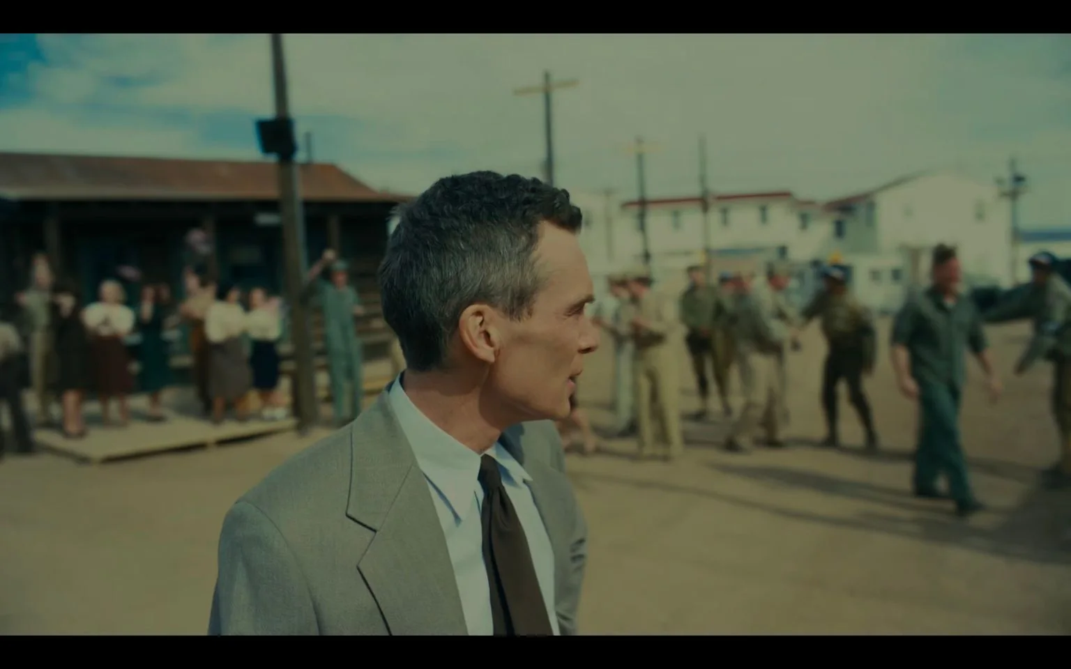 In this image, Cillian Murphy is prominently featured in an outdoor setting, portraying Oppenheimer. He is dressed in a light gray suit with a white shirt and a dark tie, facing to the side, suggesting a moment of contemplation or urgency. The scene is set in what appears to be a historical context, likely a mid-20th-century military or scientific community, as indicated by the presence of uniformed individuals and period-appropriate buildings in the background.
The atmosphere is dynamic, with several people in the background engaging in various activities. Some are in military uniforms, possibly indicating a military base or scientific facility. The lighting suggests it is daytime with clear weather, adding to the historical ambiance. The composition captures a sense of movement and interaction among the groups, with people appearing to converse or possibly celebrate something significant. The image evokes a mood of anticipation and intensity, reflecting the historical importance associated with Oppenheimer's narrative.