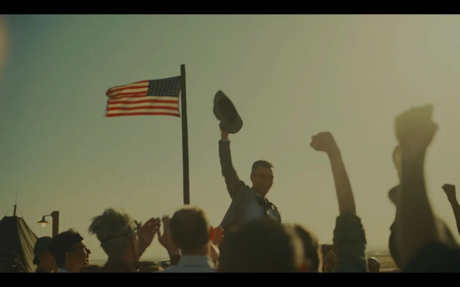 Cillian Murphy is depicted in an outdoor scene, portraying a moment from the film "Oppenheimer." He is shown triumphantly holding a hat in the air, surrounded by a crowd of people with raised fists, suggesting a celebratory or victorious atmosphere. The American flag is prominently flying in the background, adding a sense of patriotism to the scene. The lighting is warm and bright, likely capturing the essence of a sunny day. Murphy appears to be smiling, embracing the mood of the occasion. The crowd's clothing and the presence of a tent suggest a historical setting, possibly mid-20th century. The color palette is warm, dominated by shades of beige and blue, contributing to the nostalgic feel of the scene.