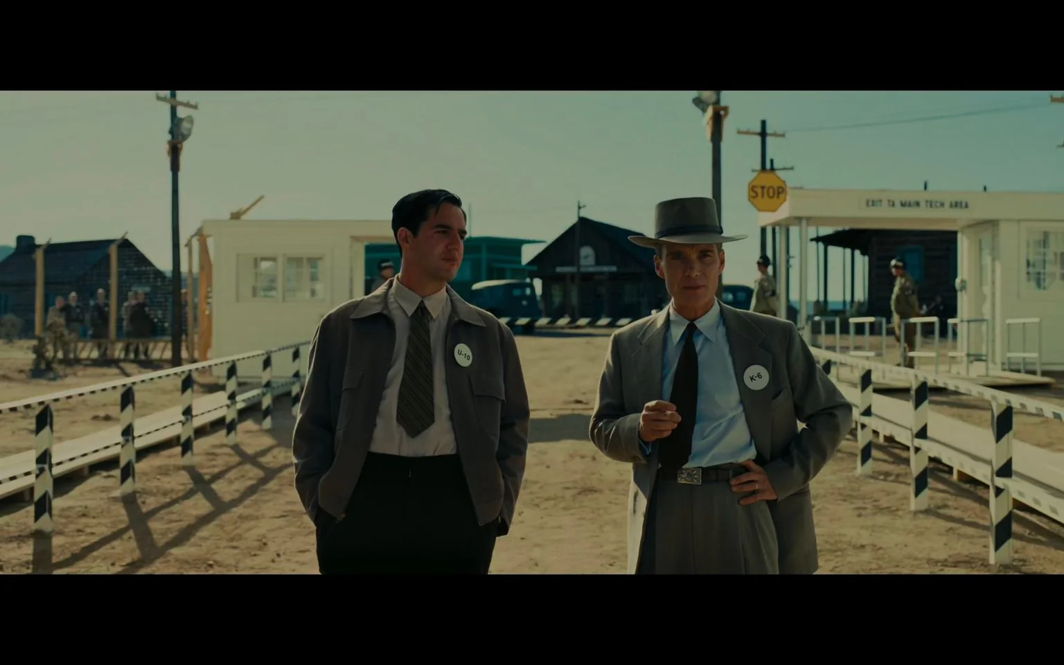 In this scene from the film "Oppenheimer," Cillian Murphy is prominently featured. The setting is an outdoor, daytime scene at what appears to be a military checkpoint. Murphy, dressed in a classic 1940s-style suit and hat, is holding a cigarette in one hand. His attire, including a light gray suit, crisp white shirt, and dark tie, reflects the period's fashion. A noticeable detail is the badge labeled "K-6" pinned to his lapel.
Beside Murphy is another man wearing a darker jacket with a badge labeled "U-10." He has a more casual posture, with his hands in his pockets. The background shows a guarded area with soldiers and a sign reading "EXIT TO MAIN TECH AREA," indicating a secure, possibly restricted location. The lighting is natural, suggesting a bright, sunny day, enhancing the scene's period authenticity and tension. The composition effectively uses depth to show the checkpoint's structure and military presence, setting a serious and historical tone.