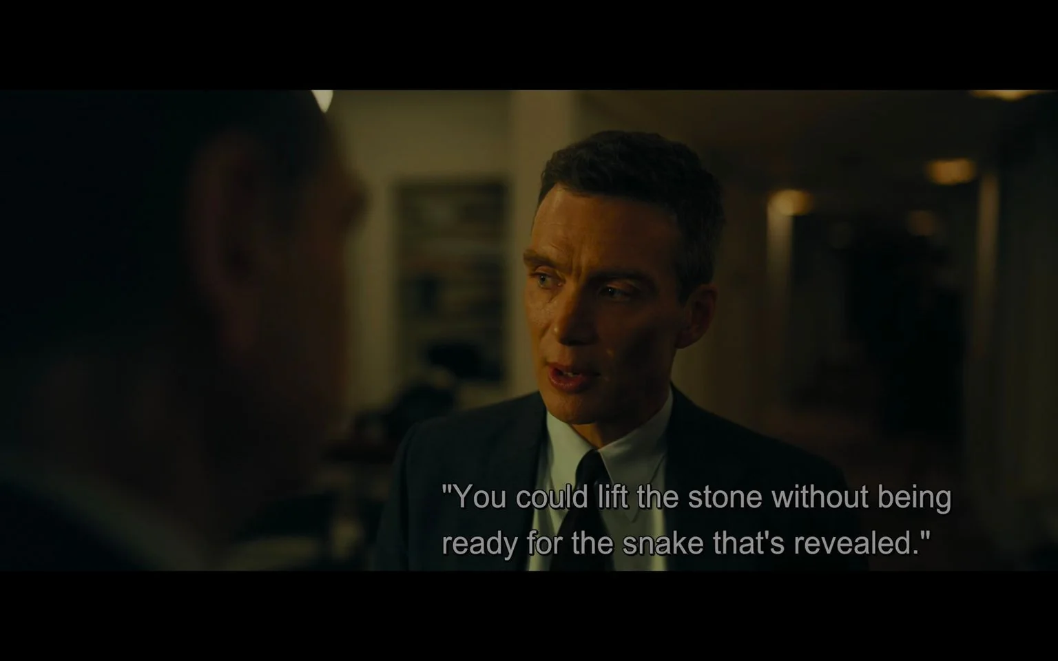 Cillian Murphy is featured in this scene from the film "Oppenheimer." The setting is indoors with subdued, warm lighting that creates a serious and intense atmosphere. Murphy is dressed in a formal dark suit and tie, suggesting a professional or high-stakes environment. His expression is focused and thoughtful, conveying deep contemplation or concern as he speaks. The background is softly blurred, indicating a room with minimal distractions, possibly an office or a meeting space. The scene captures a moment of dialogue, with the subtitle displaying, "You could lift the stone without being ready for the snake that's revealed." This metaphorical statement hints at underlying tensions or unexpected consequences in the storyline. The framing centers Murphy prominently, emphasizing the gravity of the conversation.
