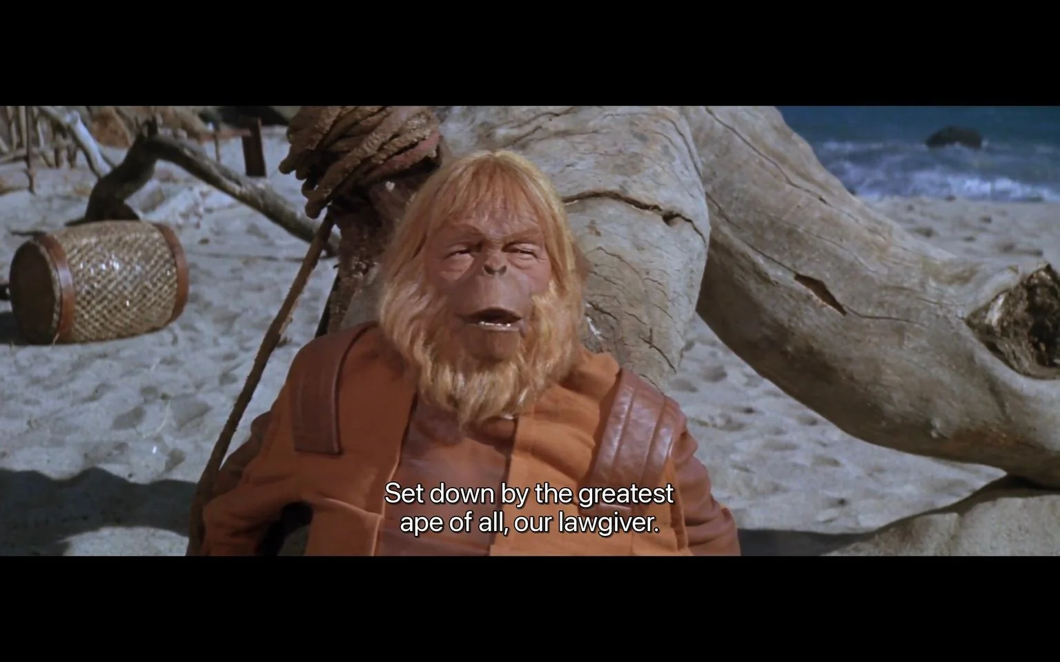 In this image from the film "Planet of the Apes," a humanoid ape character is prominently featured on a sandy beach. The setting is outdoors, during the daytime, and includes a backdrop of driftwood and the ocean. The character has long, light brown hair and a beard, wearing a brown leather-like garment, which adds to the film's science fiction aesthetic. The expression on the character’s face appears to be serious, as if delivering an important statement. The subtitle at the bottom of the image reads, "Set down by the greatest ape of all, our lawgiver," indicating a moment of dialogue that suggests a reference to a significant figure within the film's narrative. The composition focuses on the character, with objects like a rope and a woven basket visible in the background, enhancing the scene's immersive quality.