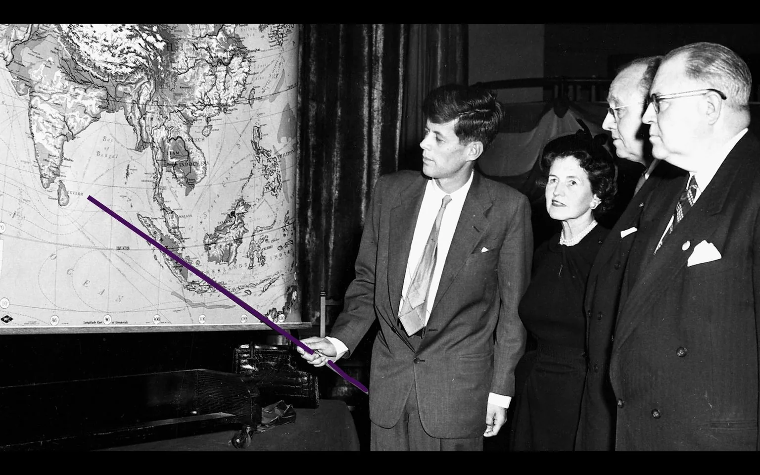 JFK is seen in this historical black and white photograph, engaging with a group of individuals in what appears to be a formal setting. He's dressed in a suit and tie, presenting with a focused expression and holding a pointer to a large map. The map prominently displays regions of Asia, including India and Southeast Asia, suggesting a discussion on geopolitical or global matters. The other individuals, also in formal attire, are attentively observing the map, suggesting a serious and possibly strategic conversation. The setting is indoors, with a curtain and what seems to be a desk or table in the foreground. The mood is formal and contemplative, highlighted by the attire and the map, which serves as the primary focal point of the discussion.