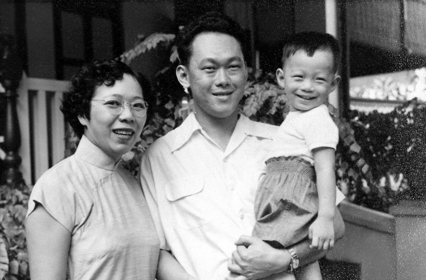 In this black and white vintage photograph, a family portrait captures Lee Kuan Yew holding a young child, Lee Hsien Loong. The setting appears to be outdoors, with a backdrop of foliage and a partial view of a porch railing. Lee Kuan Yew is wearing a light-colored, short-sleeved shirt, and he has a gentle smile on his face. He cradles Lee Hsien Loong, who is a small child at the time, wearing a light shirt and knee-length shorts, smiling joyfully at the camera. To the left, a woman stands beside them, dressed in a traditional high-collared blouse, smiling warmly. The overall mood is one of familial warmth and affection, capturing a personal moment in the early years of a prominent family. The image exudes a sense of nostalgia, typical of family photos from mid-20th century Singapore.
