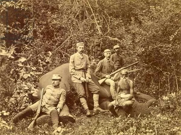 The image captures a historical scene featuring Archduke Franz Ferdinand and a group of men posing with a large game animal in a forested area. Five individuals are present, dressed in attire typical of the late 19th to early 20th century, with some wearing military-style uniforms and caps. The group is gathered around the carcass of a large animal, likely hunted during an expedition.
Archduke Franz Ferdinand is among the group, identifiable from historical context rather than visual recognition. The men are armed with rifles, which they hold or rest on the ground. The setting is dense with foliage, indicating a jungle or forested environment, and the lighting suggests a daytime scene with natural light filtering through the trees. The image has a sepia tone, common in photographs from this era, adding to its vintage quality. The expressions and posture of the men convey a sense of accomplishment and formality typical of such hunting excursions during that period.