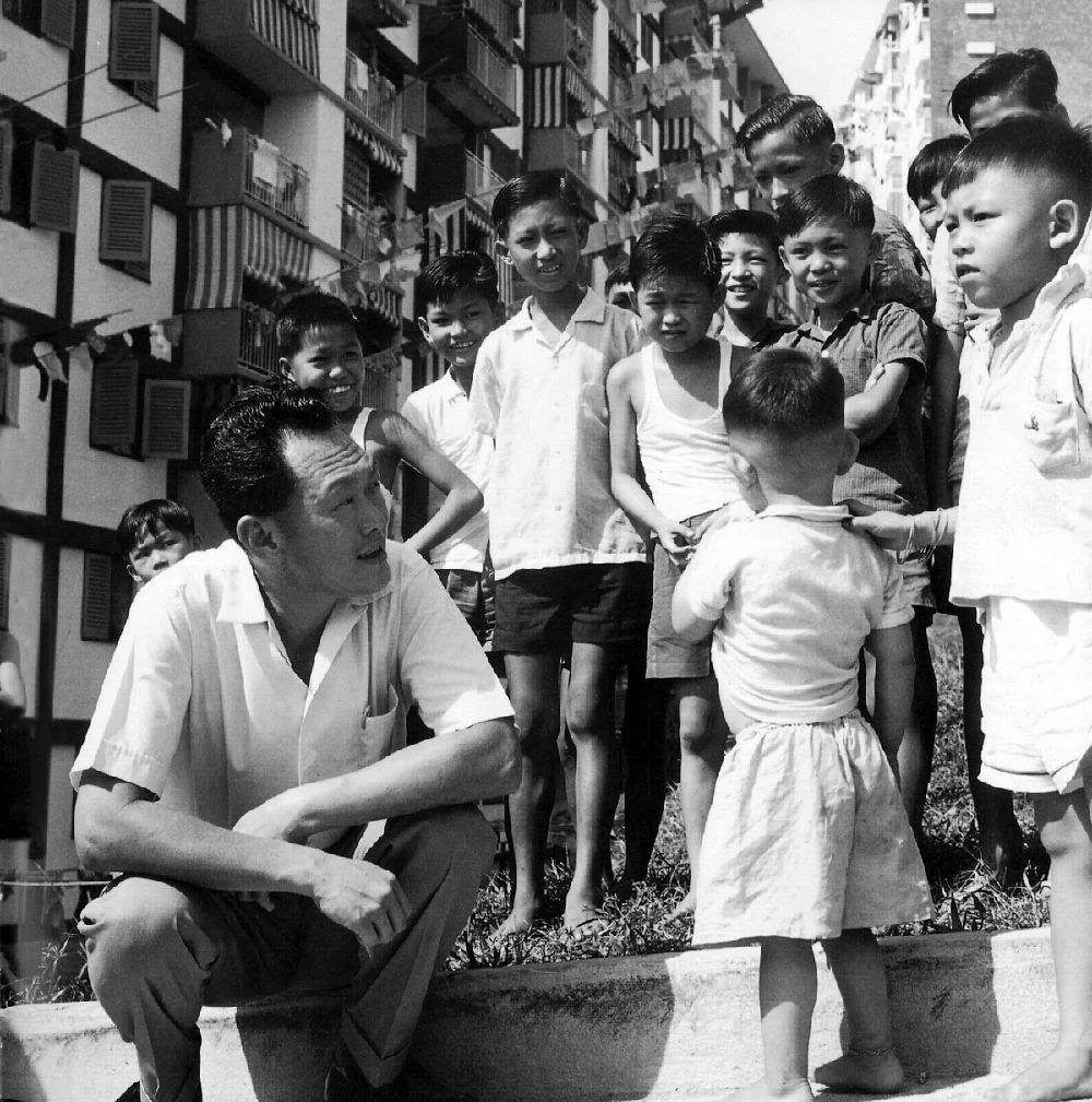 In this black-and-white photograph, Lee Kuan Yew is seen interacting with a group of children. He is crouching down, wearing a light-colored short-sleeved shirt and pants, appearing engaged and focused on the children surrounding him. The children, who are smiling and appearing curious, are dressed in casual, everyday clothing typical of the era, such as shorts and simple shirts. They are gathered closely around Lee Kuan Yew, emphasizing a sense of community and attentiveness.
The background shows a series of residential buildings, suggesting an urban setting. The architecture appears to be mid-20th century, with visible balconies and open windows typical of the time. The lighting suggests it is daytime, with the sun casting shadows and highlighting the expressions on the children’s faces. The overall mood is one of warmth and connection, capturing a moment of interaction and engagement between a leader and the younger generation.