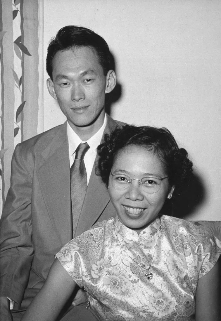 The image is a black-and-white portrait featuring Lee Kuan Yew alongside a woman, likely in a formal setting. Lee Kuan Yew is dressed in a suit with a tie, exuding a composed and confident demeanor. The woman is wearing a traditional, patterned dress with short sleeves and glasses, smiling warmly at the camera. They are seated closely together, suggesting a sense of intimacy or partnership. The background shows a curtain with a leaf pattern, indicating an indoor setting. The lighting is even, typical of portraiture from the mid-20th century, highlighting their features without harsh shadows. The overall mood is formal yet relaxed, capturing a moment of personal significance.