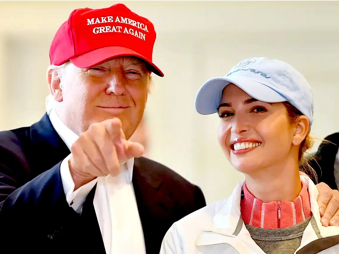 President Trump is standing next to a woman in this indoor setting. He is wearing a bright red baseball cap with the slogan "MAKE AMERICA GREAT AGAIN" prominently displayed in white letters. His attire includes a dark suit jacket over a white shirt, and he appears to be in a relaxed and jovial mood, smiling and pointing towards the camera. Beside him is a woman wearing a light blue baseball cap and a casual outfit that includes a white jacket and a pink turtleneck. She is smiling warmly, adding to the friendly and informal atmosphere of the scene. The lighting is bright and even, contributing to the cheerful ambiance. The image captures a moment of camaraderie between the two individuals.