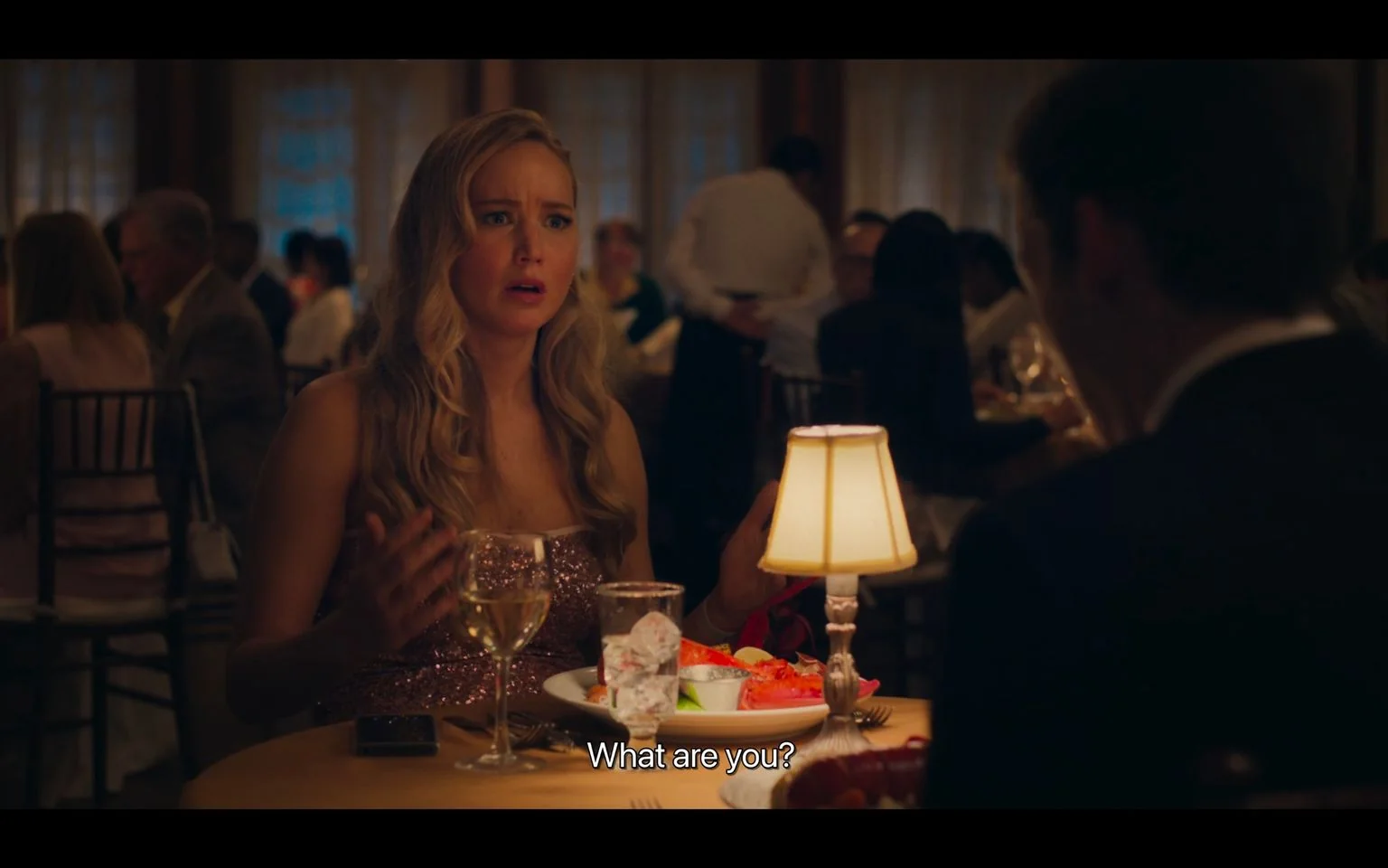 The image captures a scene set in a dimly lit restaurant, creating an intimate and sophisticated ambiance. A woman, wearing a sparkling dress, is seated at a dining table. Her facial expression is one of surprise or confusion, suggesting a moment of intense dialogue. In front of her is a glass of white wine, a plate with food, and a smartphone on the table. The table is elegantly set, with a small lamp casting a warm glow over the scene. In the background, other patrons are seen engaged in conversation, enhancing the bustling yet private feel of the restaurant. The subtitles at the bottom read, "What are you?" indicating a pivotal or dramatic moment in their conversation. The overall atmosphere is one of elegance and tension, with the woman's expressive demeanor suggesting a crucial point in the narrative.