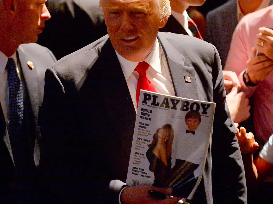 The image depicts President Trump in an indoor setting, surrounded by a crowd. He is wearing a dark suit with a white shirt and a red tie, complemented by a small U.S. flag pin on his lapel, which is a typical accessory for public appearances. The lighting appears somewhat dim, casting shadows on his face, but his distinctive hairstyle and confident demeanor are noticeable.
President Trump is holding a "Playboy" magazine prominently in his hand. The magazine's cover features the title "PLAYBOY," with the subtitle "ENTERTAINMENT FOR MEN" beneath it. The cover includes a headline about a "DONALD TRUMP INTERVIEW," suggesting this edition features content related to him. There is an image of a woman in a dress and a man in a tuxedo on the cover, likely related to the interview or feature article.
The scene seems to capture a candid moment, potentially at a public or promotional event, with several people around, some engaged in conversation. The atmosphere appears lively, with individuals in professional attire, indicating a formal or semi-formal occasion.