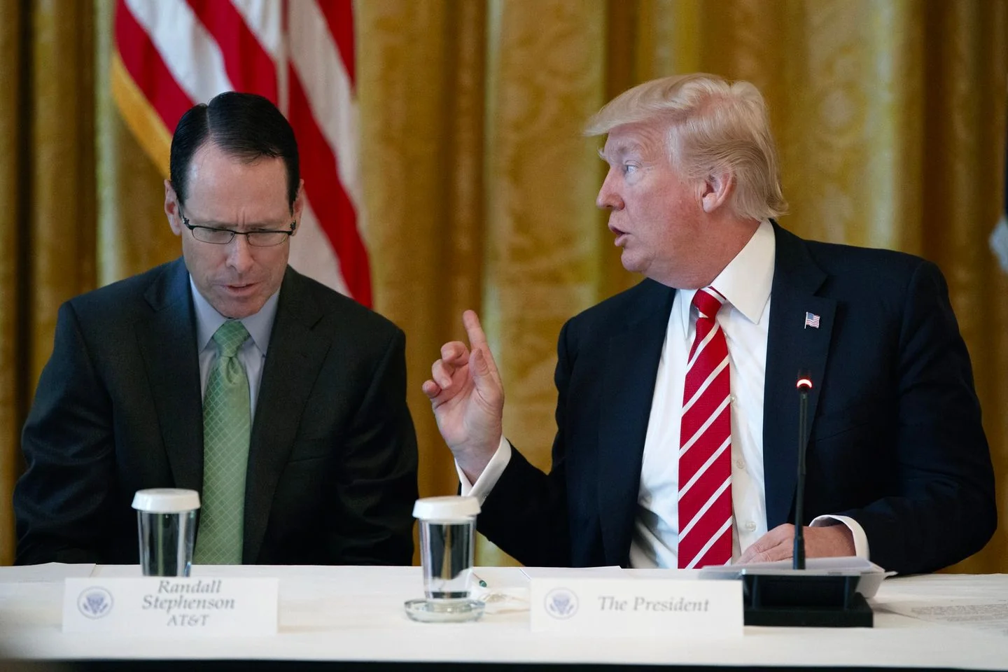 The image captures a formal meeting setting featuring two individuals seated at a long table, engaged in a discussion. On the right is "The President," who is speaking and gesturing with his right hand. He is wearing a dark suit with a white shirt and a red and white striped tie, complemented by a small American flag pin on his lapel. His expression appears focused and conversational.
To his left is Randall Stephenson, identified by the nameplate in front of him, along with his affiliation with AT&T. He is wearing a dark suit, a light blue shirt, and a green tie, appearing to be listening intently. Both individuals are seated in front of a gold-colored curtain, with part of an American flag visible in the background. There are microphones and glasses of water on the table, indicating a formal discussion or conference setting. The lighting is bright, highlighting the serious and professional atmosphere of the scene.
- Title: "..."
- Channel / profile: "..."
- Site / app: "..."