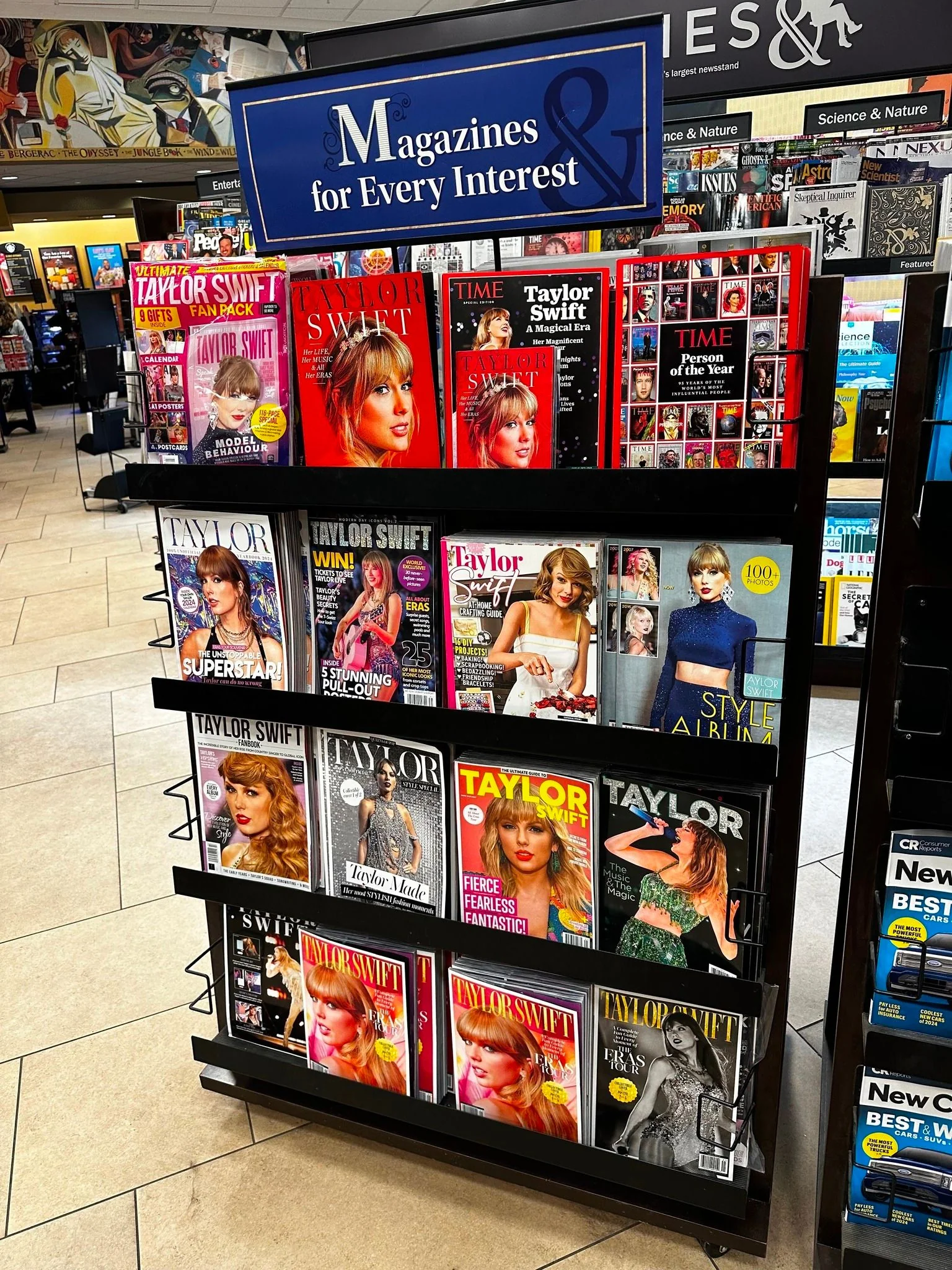 The image depicts a magazine stand indoors, prominently featuring a variety of magazines with Taylor Swift on the covers. The display is topped with a sign reading "Magazines for Every Interest" in white text on a blue background. The magazines showcase different images and styles of Taylor Swift, including several issues with her on the cover of "TIME" magazine, notably one titled "TIME Person of the Year." The covers are vibrant, with bright colors like reds, yellows, and blues, highlighting various aspects of her career and style. Taylor Swift appears in multiple outfits and poses across the covers, with themes ranging from music to fashion. The background of the image shows a store setting with more magazine stands and a mural on the wall. The lighting is bright, typical of a retail environment, enhancing the vividness of the magazine covers.