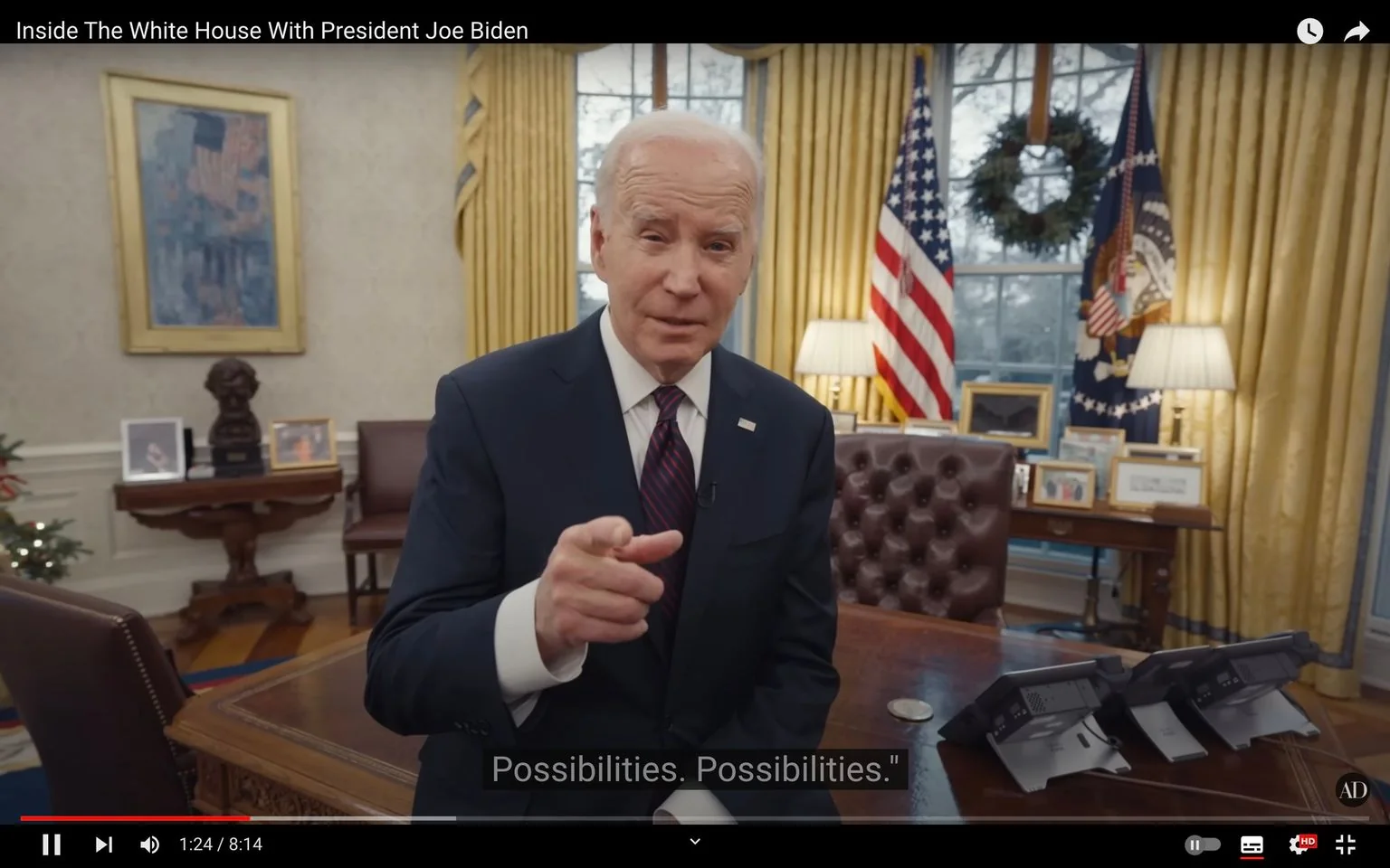 The video frame shows President Joe Biden speaking inside the Oval Office. He is pointing towards the camera, suggesting an engaging and direct communication. The setting is formal and professional, characterized by the classic decor of the White House. Behind him, the American flag and the presidential flag are prominently displayed, along with a wreath hanging in the window, hinting at a holiday season. The room is well-lit with a warm ambiance, courtesy of the natural light through the window and table lamps. On his desk, several digital devices are visible, alongside framed photographs and documents, indicating a working environment. The furniture includes a tufted leather chair and a wooden desk, enhancing the traditional feel. The video title is "Inside The White House With President Joe Biden," indicating the context of a tour or interview. Subtitles in the image capture him saying the word "Possibilities" twice, underlining a theme of potential and hope.