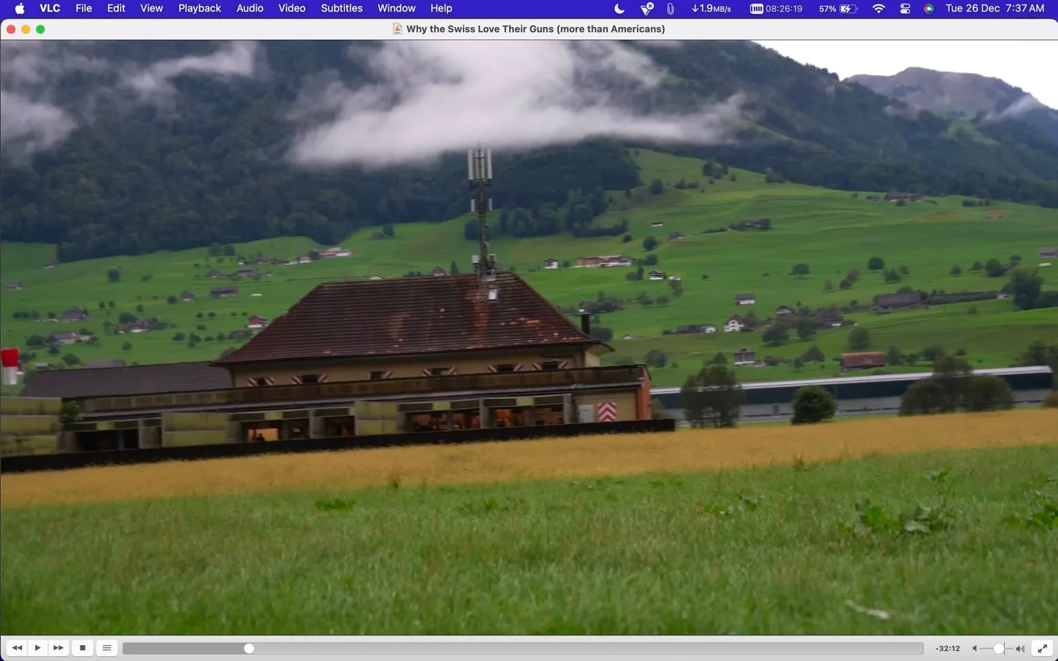 The image is a screenshot from a VLC media player showing a scene titled "Why the Swiss Love Their Guns (more than Americans)." The scene captures a picturesque rural landscape in Switzerland. In the foreground, there is a traditional Swiss building with a sloped roof and a tall antenna, possibly indicating some form of communication equipment. Surrounding this building are expansive green fields, which reflect the lushness typical of the Swiss countryside.
In the background, the image displays rolling hills dotted with small houses, suggesting a sparsely populated rural area. A layer of low-lying clouds drapes over the distant hills, adding a dramatic and serene atmosphere to the setting. The overall lighting is natural, implying that the scene was captured during the daytime. No visible people or movement can be seen, emphasizing the tranquility of the rural environment. The VLC interface displays standard playback controls and indicates that the content is paused.