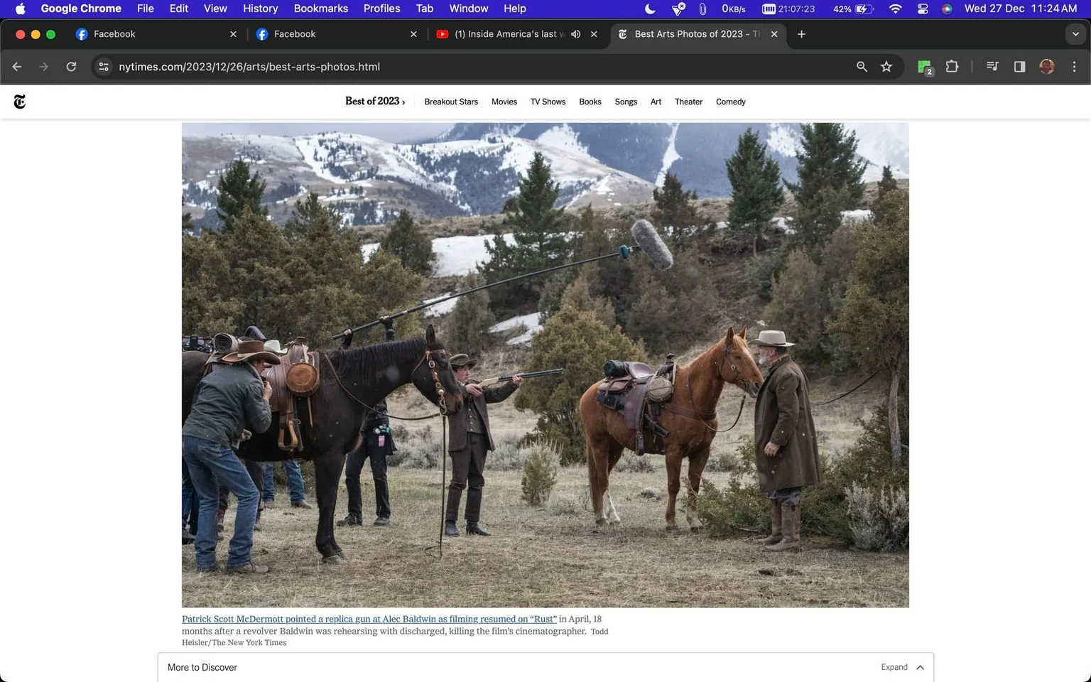 The image depicts a film set for the movie "Rust" with cast and crew members engaged in a scene. The setting is a rugged, mountainous landscape with patches of snow visible on the hills in the background, suggesting a winter environment. The scene is outdoors, surrounded by trees and uneven terrain. In the foreground, two horses are saddled, indicative of a western film theme. Several individuals are present, including Patrick Scott McDermott, who is holding a replica gun pointed at Alec Baldwin. Alec Baldwin, wearing a long coat and hat, stands near one of the horses. A boom microphone is visible above the group, capturing audio for the scene. The lighting appears natural, consistent with the outdoor setting. The scene is part of the film's production resumption following a previous incident involving a firearm on set. This image is part of an article titled "Best Arts Photos of 2023" published by The New York Times.
- Title: "Best Arts Photos of 2023"
- Site / app: "nytimes.com"
- Captions / subtitles: "Patrick Scott McDermott pointed a replica gun at Alec Baldwin as filming resumed on “Rust” in April, 18 months after a revolver Baldwin was rehearsing with discharged, killing the film’s cinematographer. Todd Heisler/The New York Times"