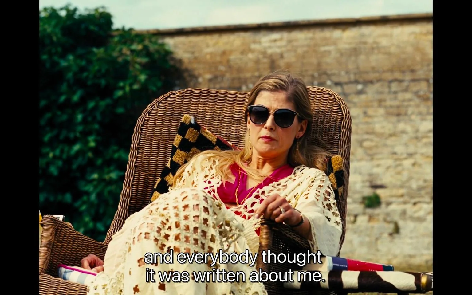 The scene captures an outdoor setting, featuring a person lounging in a wicker chair. The individual is wearing sunglasses and is draped in a crocheted shawl, suggesting a relaxed and casual atmosphere. They appear to be in a contemplative mood, with a subtle expression on their face as they speak. The background includes a stone wall and lush greenery, adding a serene and picturesque touch to the environment.
The lighting is warm and natural, indicating it might be a sunny day, which complements the laid-back vibe of the scene. The individual is dressed in a vibrant pink top, adding a pop of color against the neutral tones of the shawl and chair. In the foreground, a checkered blanket is visible, further enhancing the cozy atmosphere.
The caption on the image reads, "and everybody thought it was written about me," suggesting that the person is reflecting on a personal or public perception regarding something significant to them.