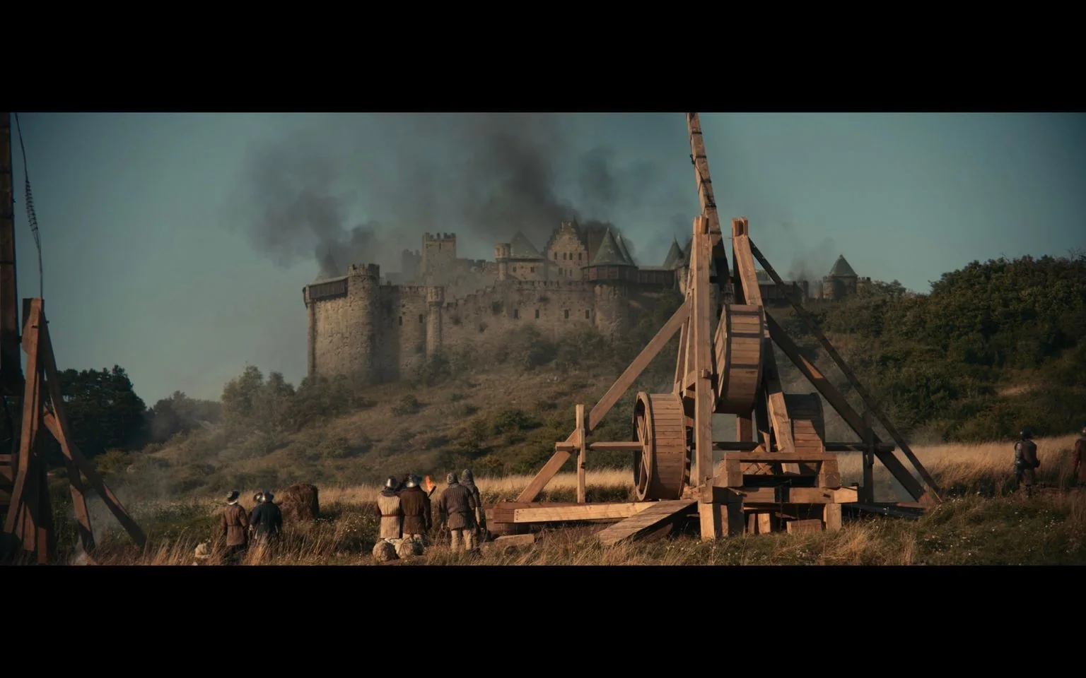 The image portrays a medieval siege scene, likely from "Kingdom Come: Deliverance." In the foreground, a large wooden trebuchet, a historical siege weapon, is prominently displayed, ready to launch projectiles. Several figures in period-appropriate attire, including helmets and tunics, are gathered around the trebuchet, possibly soldiers preparing for an assault. In the background, a formidable stone castle sits atop a hill, surrounded by lush greenery. Dark smoke billows from the castle, indicating ongoing battle or destruction.
The lighting suggests a clear day, with natural sunlight illuminating both the trebuchet and the castle. The overall mood is tense and dramatic, capturing the intensity of a siege. The scene's composition balances the human figures, the powerful machinery of war, and the distant castle, emphasizing the scale and gravity of the conflict. The style is realistic, aligning with the historical setting of the game.