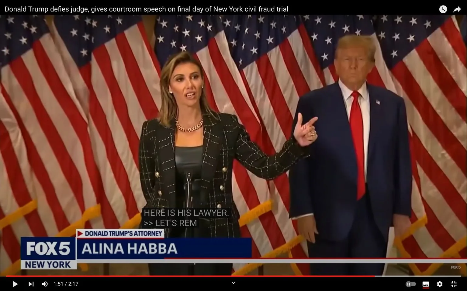Alina Habba is seen speaking in front of a backdrop of American flags, indicating a formal setting such as a press conference. She is dressed in a black, patterned blazer with a gold chain necklace, exuding a professional appearance. Her expression is focused, and she gestures with her hand, possibly emphasizing a point. Standing beside her is a man in a navy suit with a red tie, maintaining a serious demeanor.
The setting is likely a formal or legal event, as suggested by the presence of multiple flags and the attire of the individuals. The lighting is bright, highlighting the two figures. Text on the screen identifies Alina Habba as "Donald Trump's Attorney," beneath the FOX 5 New York branding, indicating a news broadcast. Captions on the screen read, "HERE IS HIS LAWYER," followed by an incomplete line, "LET'S REM," suggesting a continuation of a discussion or statement. The title text provides context about the event, mentioning a courtroom speech by Donald Trump.