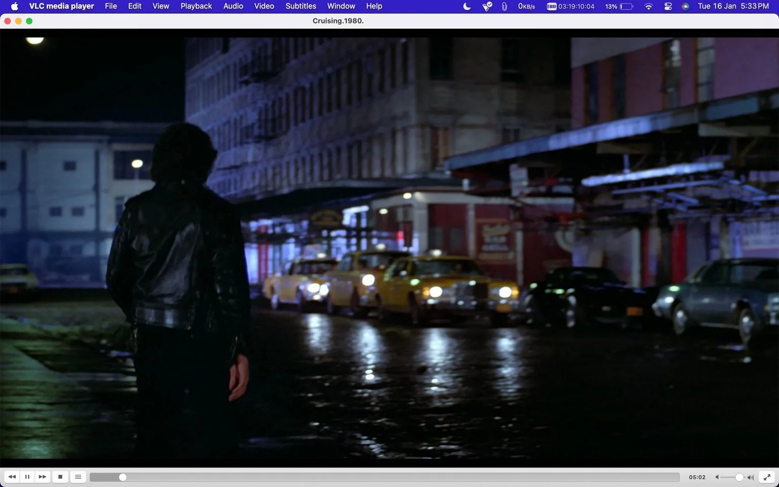 In this screenshot from the film "Cruising" (1980), we see a scene set at night in an urban environment. The focus is on a character, portrayed by Al Pacino, who is seen from behind. He wears a black leather jacket, adding to the mysterious and gritty atmosphere typical of a thriller. The character is walking down a wet street, reflecting the lights from the surrounding buildings and vehicles, which include several yellow taxis parked along the side.
The lighting is dim, enhancing the noir feel, with the streetlights and building signs providing the primary illumination. The scene captures the essence of a bustling city at night, with the architecture of the buildings and the parked cars contributing to the setting's authenticity. The mood is tense and immersive, drawing the viewer into the narrative's suspenseful world. The use of a VLC media player interface suggests this is a digital viewing, with familiar playback controls and menu options visible at the top and bottom of the screen.