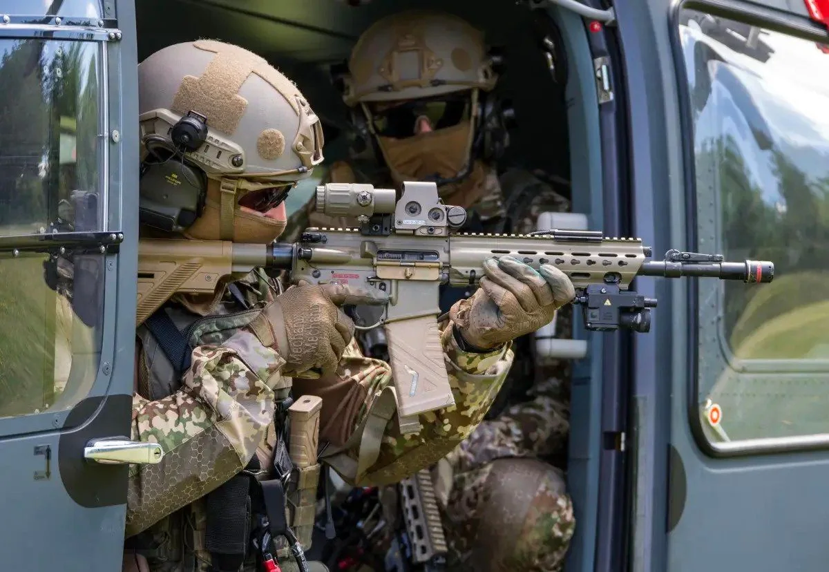 The image depicts two soldiers in full tactical gear inside a helicopter. The soldier in the foreground is aiming a rifle out of the helicopter's open door. They are wearing camouflage uniforms, tactical gloves, and helmets equipped with headsets and goggles. The rifle is fitted with various attachments, including a scope and a foregrip. The other soldier, partially visible in the background, is similarly equipped and appears to be observing the surroundings. The helicopter's interior is visible, with the door open to a view of greenery outside, suggesting the helicopter is either in flight or stationary close to the ground. The scene conveys a sense of readiness and precision, with the soldiers focused on their task.