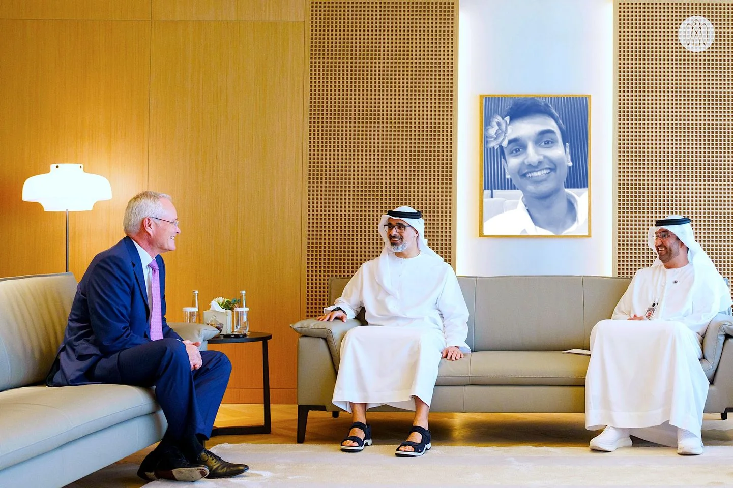 In this image, three men are seated in an elegantly designed room, engaged in a conversation. The decor features a modern aesthetic with warm wooden tones and a soft glowing lamp on the left. The man on the left wears a blue suit and a pink tie, sitting on a couch facing the two other men in traditional attire. The middle man, dressed in a white kandura and sandals, smiles as he converses with the man on the right, who is also in traditional attire and appears to be holding papers. A framed black-and-white painting of Lehan hangs prominently on the wall behind them, adding a personal touch to the setting. The atmosphere is formal yet relaxed, with elements like bottled water and a small flower arrangement on the table emphasizing the setting's hospitality. The lighting is bright and even, highlighting the clean and polished interior.