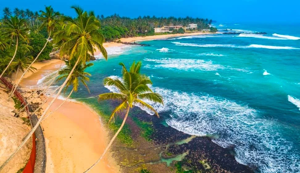 The image showcases a stunning tropical beach scene from an aerial perspective. In the foreground, several tall palm trees lean gracefully over a pristine sandy beach. The sand is a soft, golden color, blending seamlessly into the turquoise water of the ocean. Gentle waves roll in, creating a pattern of white foam as they break against the shore and the rocky areas visible beneath the clear water. In the background, more palm trees line the coastline, and a few buildings are nestled among the lush greenery, suggesting a serene and secluded setting. The sky is a vibrant blue, enhancing the overall tropical and tranquil atmosphere of the scene. The lighting is bright and natural, highlighting the vivid colors and creating a sense of warmth and paradise.