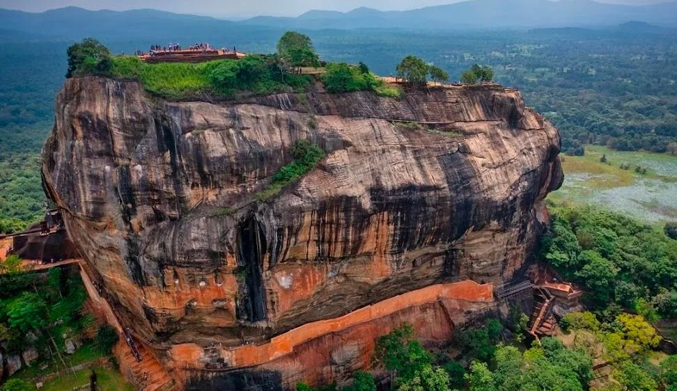 The image depicts the ancient rock fortress known as Sigiriya, located in Sri Lanka. This iconic landmark features a massive column of rock, approximately 200 meters high, rising dramatically from the lush green forest that surrounds it. The top of the rock is flat and hosts the ruins of an ancient civilization, with visible remnants of staircases and terraces leading up to it. A group of people can be seen at the summit, suggesting it's a popular tourist destination.
The rock itself has a striking appearance, with natural patterns and color variations in shades of gray, brown, and orange, giving it an imposing yet beautiful look. Trees and vegetation are scattered across the top and around the base, adding to the natural beauty of the setting. The surrounding landscape is expansive, with a vast stretch of greenery and distant hills visible in the background, evoking a sense of tranquility and grandeur. The lighting is bright and even, indicating it is likely captured during daylight, enhancing the vividness of the colors.