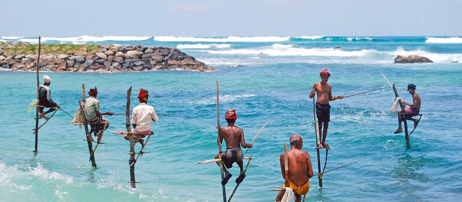 The image depicts a group of traditional fishermen practicing stilt fishing in the ocean. They are sitting on tall wooden poles planted in the shallow coastal waters, a method distinctive to Sri Lanka. This technique involves balancing on a horizontal crossbar, known as a "petta," and using long rods to catch fish. The fishermen are dressed in simple attire, with some wearing colorful head wraps. The water is a vivid turquoise, and the waves gently lap against the rocks in the background, creating a picturesque seaside setting. The bright daylight highlights the vibrant colors of the ocean and the fishermen's clothing. The scene conveys a sense of timeless tradition and harmony with nature, with the fishermen focused on their task, waiting patiently for a catch.