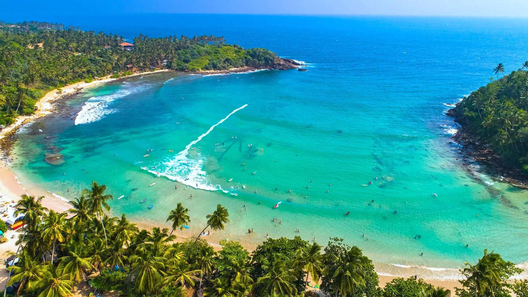 This image showcases a stunning aerial view of a tropical beach, surrounded by lush green palm trees and vibrant turquoise waters. The bay is crescent-shaped, with gentle waves lapping against the sandy shore. Numerous people can be seen enjoying the water, some appear to be swimming while others are possibly paddleboarding or engaging in other water activities. The beach is framed by dense foliage, adding to the secluded and serene atmosphere of the scene. The water is a rich gradient of blues and greens, with the waves creating a textured surface that contrasts beautifully against the smooth sand. The lighting is bright and sunny, indicating a clear day, which enhances the colors and adds a warm, inviting feel to the image. This setting evokes a sense of relaxation and tropical bliss, ideal for a beach getaway.