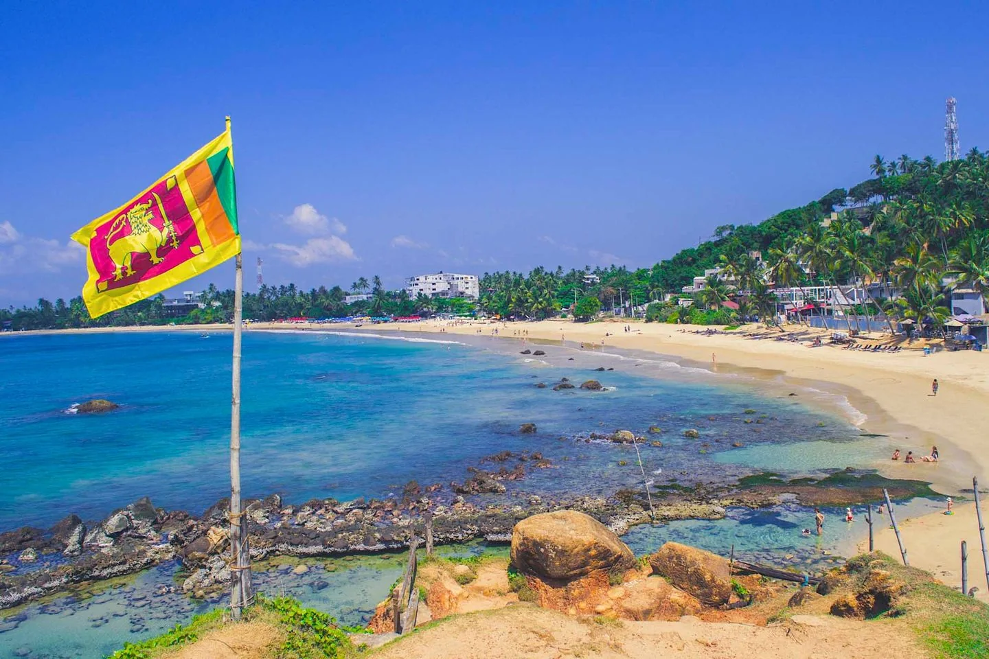 The image depicts a beautiful beach scene in Sri Lanka. In the foreground, the Sri Lankan flag is prominently displayed, fluttering in the breeze atop a wooden pole. The flag's vibrant colors of yellow, green, and red stand out against the bright blue sky and are a symbol of the nation's rich heritage. The coastline stretches out into the distance, with clear turquoise waters lapping gently against the sandy shore. Scattered rocks and corals are visible along the water's edge, adding texture to the scene.
In the background, several people are enjoying the beach, some swimming in the ocean while others relax on the sand. The lush greenery of palm trees and tropical vegetation provides a picturesque backdrop, while white buildings are partially visible among the trees, likely beachside resorts or residences. The lighting is bright and natural, suggesting a sunny day, perfect for beach activities. The overall mood of the image is serene and inviting, capturing the essence of a tropical paradise.