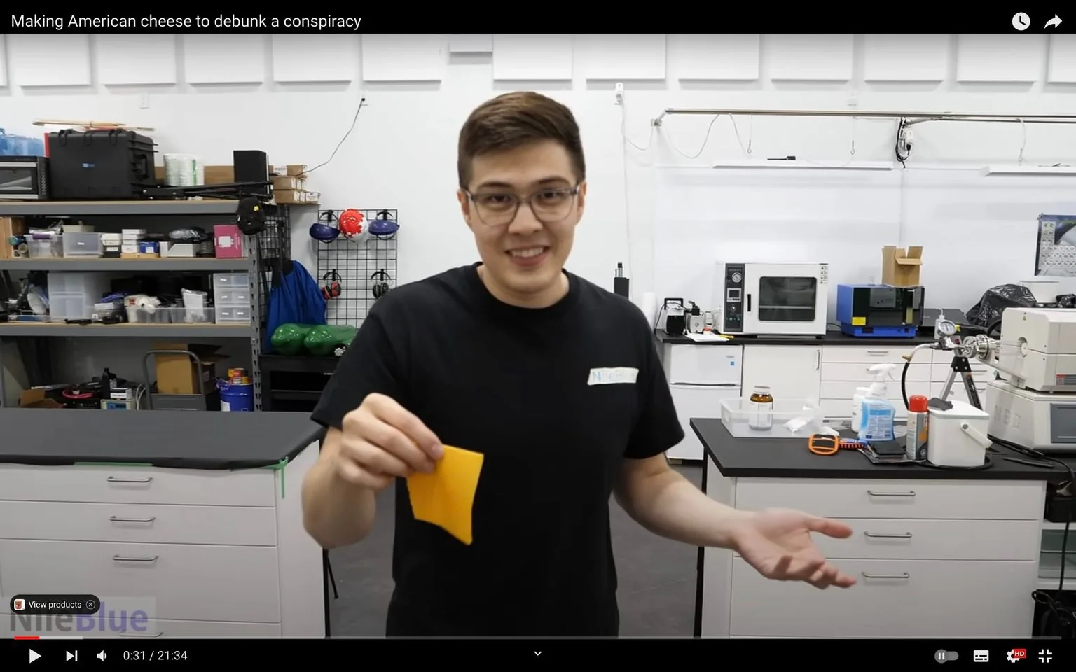 The image is a video frame from a video titled "Making American cheese to debunk a conspiracy" on the NileRed channel. The scene is set indoors in a laboratory, suggesting an experimental or scientific environment. The person in the frame is holding a slice of orange cheese and appears to be explaining or demonstrating something. He is wearing a black T-shirt with a name tag reading "NileBlue," indicative of branding or channel affiliation.
The laboratory is well-equipped, with shelves containing various items such as boxes and equipment, and there are workbenches with scientific devices and containers. The background includes storage shelves, a pegboard with tools and safety gear, and equipment like an oven and other lab apparatus. The person has a friendly demeanor, smiling while engaging with the audience, likely explaining the process involved in the experiment regarding American cheese. The lighting is bright and even, ensuring clear visibility of the details in the lab and the subject.