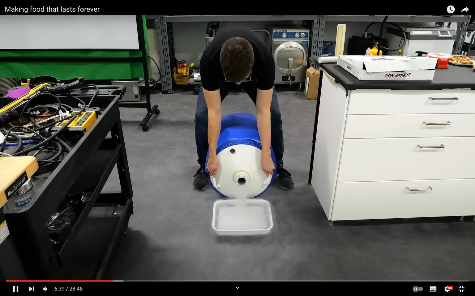 The image is a frame from a YouTube video titled "Making food that lasts forever." It shows a person in a workshop setting performing an experiment. The individual is bending down and holding a large blue and white container that appears to be dispensing a substance into a rectangular tray on the floor, creating a foggy effect around the area. This suggests the use of liquid nitrogen or a similar substance, commonly used in food preservation experiments.
The workspace is equipped with various tools and equipment. A table on the left holds numerous cables and devices, while a countertop on the right displays a pizza box labeled "Bon Appetit," along with other food items. The background features metal shelving with additional equipment, including what looks like vacuum chambers or freeze dryers, often used in preservation processes. The setting is well-lit, highlighting the scientific and experimental nature of the scene.