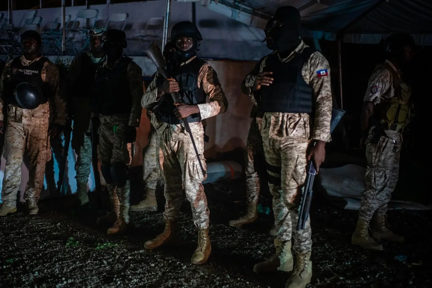 The image depicts a group of security personnel standing together at night. They are wearing camouflage uniforms and tactical gear, including helmets and vests. One officer is holding a rifle at the ready, while others have weapons or shields. The lighting is dim, suggesting a nighttime setting, with some artificial light casting shadows and highlights on their uniforms. The mood is serious and vigilant, as the officers appear to be on duty or preparing for an operation. The background includes a tent and folding chairs, adding to the impression of a temporary setup, possibly for an event or operation. The text "POLICE NATIONALE D'HAITI" is visible on one of the uniforms, indicating the presence of Haitian national police.