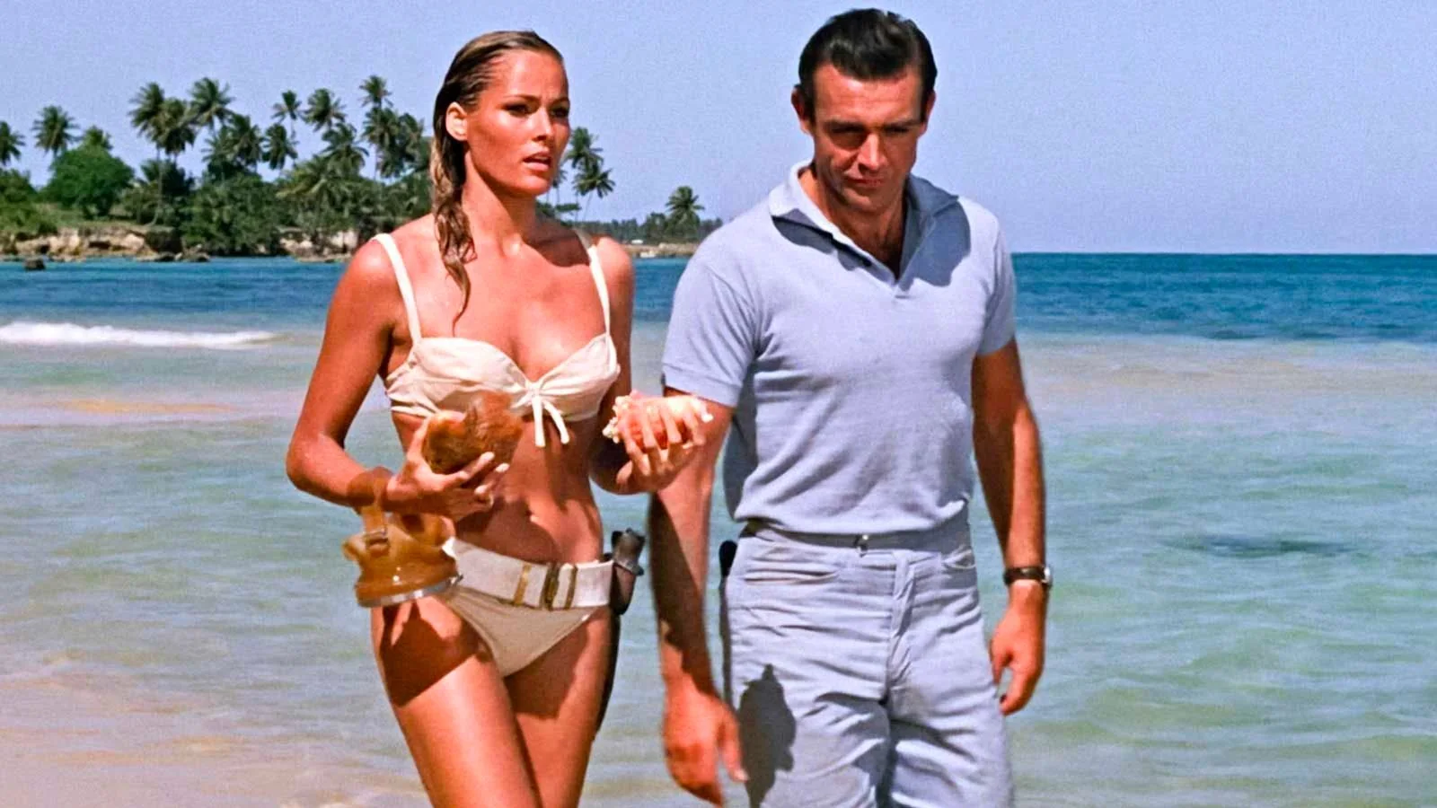 In this iconic scene from the film "Dr. No," Sean Connery is featured walking on a tropical beach. The setting is bright and sunny, with a clear blue sky and ocean in the background, creating a serene atmosphere. Sean Connery is dressed casually in a light blue polo shirt and matching trousers, exuding a relaxed yet suave demeanor typical of his portrayal of James Bond. Beside him, a woman in a white bikini is holding seashells, adding to the classic beach aesthetic. The shoreline is lined with palm trees, enhancing the tropical vibe. Both characters appear engaged in conversation, with the woman looking intently ahead while Sean Connery maintains a confident composure. The lighting is natural, highlighting the vivid colors of the ocean and the characters' attire, contributing to the vibrant, summery mood of the scene.